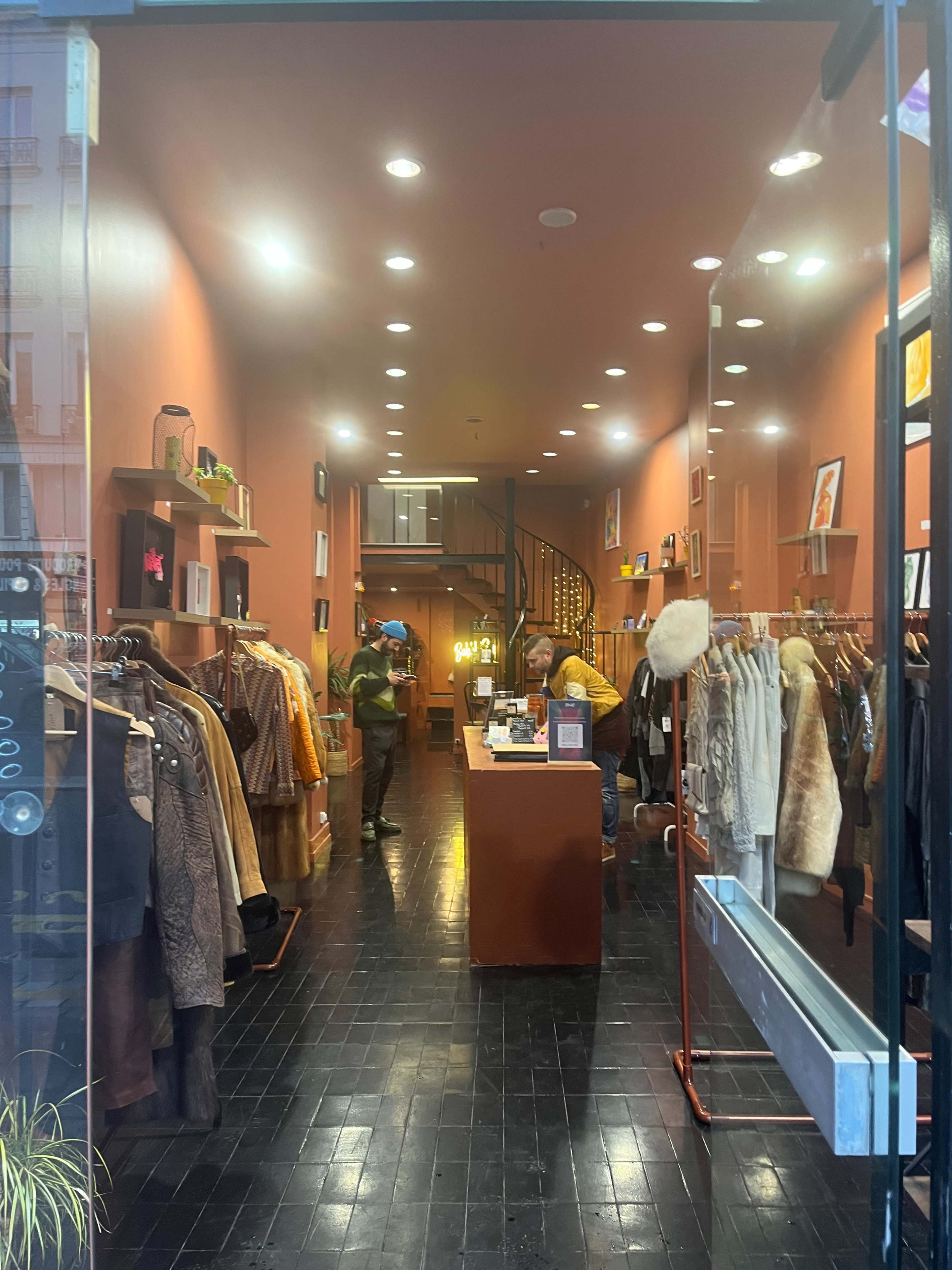 A vintage clothing store features racks of clothes and a winding staircase in the background, with customers browsing and an employee at the counter.