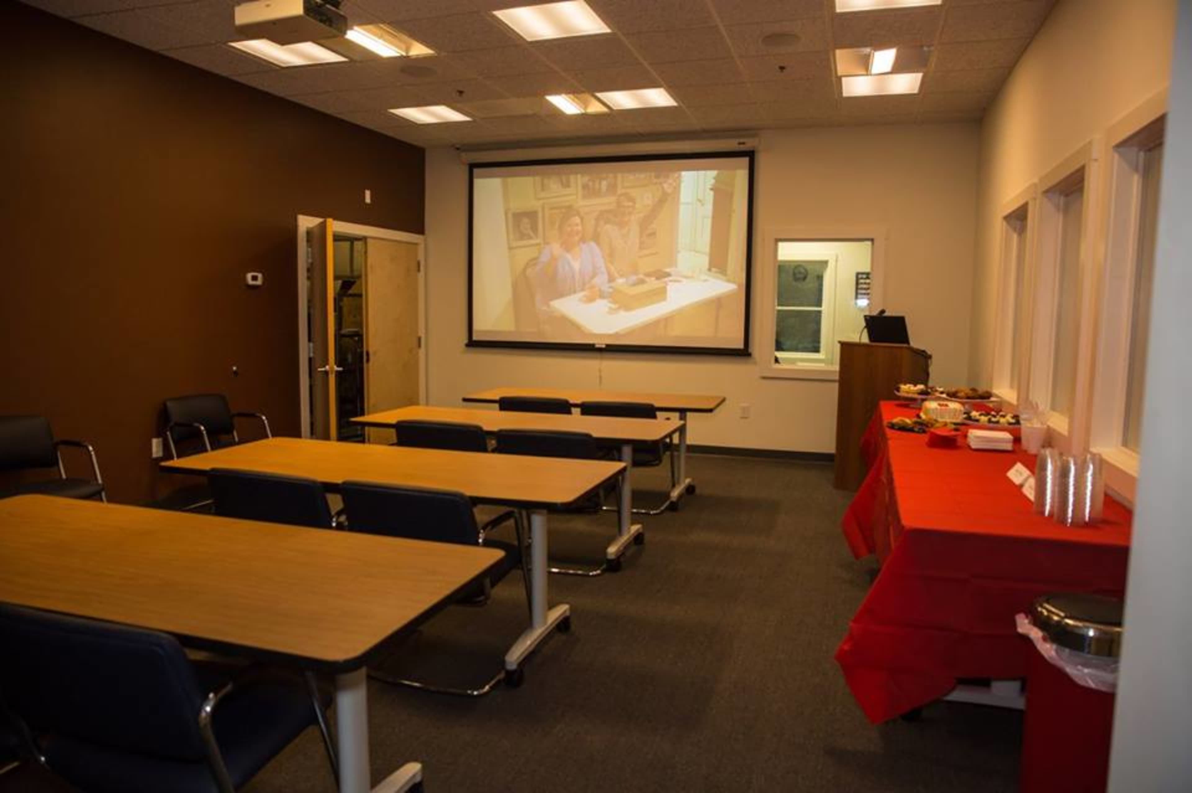 A conference room is set up with several tables and chairs facing a projected screen, alongside a food table covered with a red cloth.