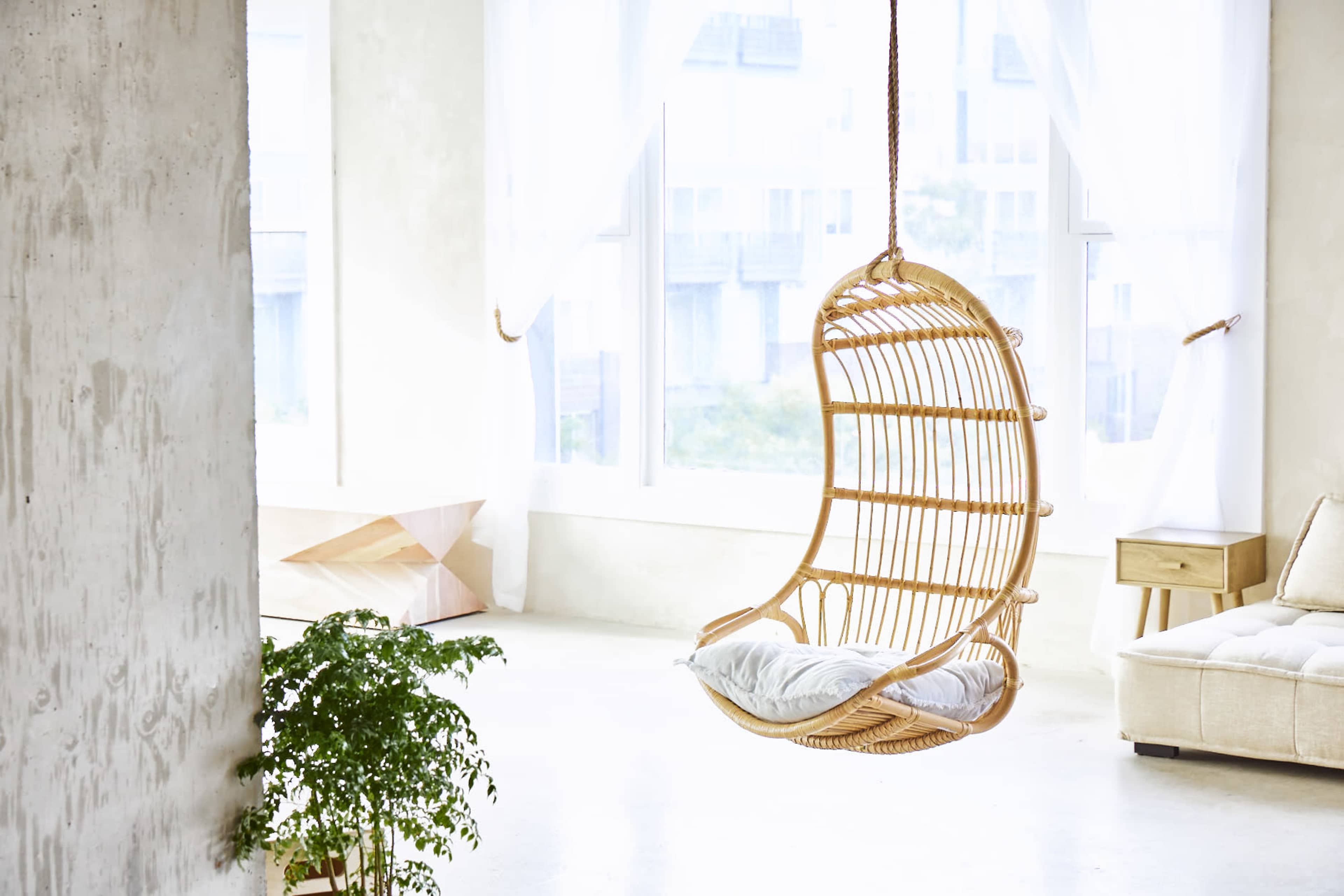 A rattan hanging chair suspended in a bright, minimalist room with large windows and a potted plant.