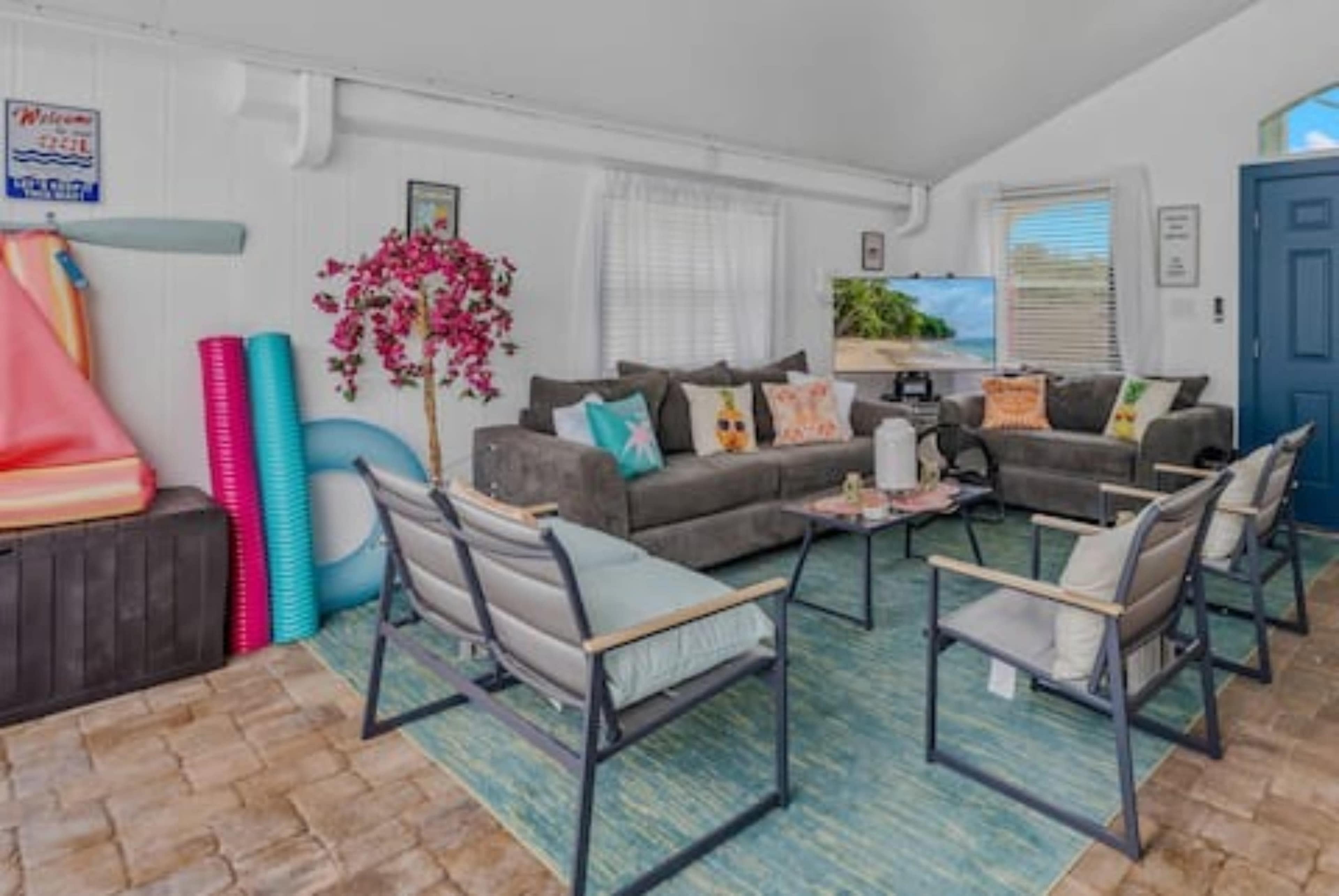 The image shows a bright and airy living room with a gray sofa, two armchairs, and a coffee table, surrounded by colorful decor and outdoor items like pool floats and a storage box.