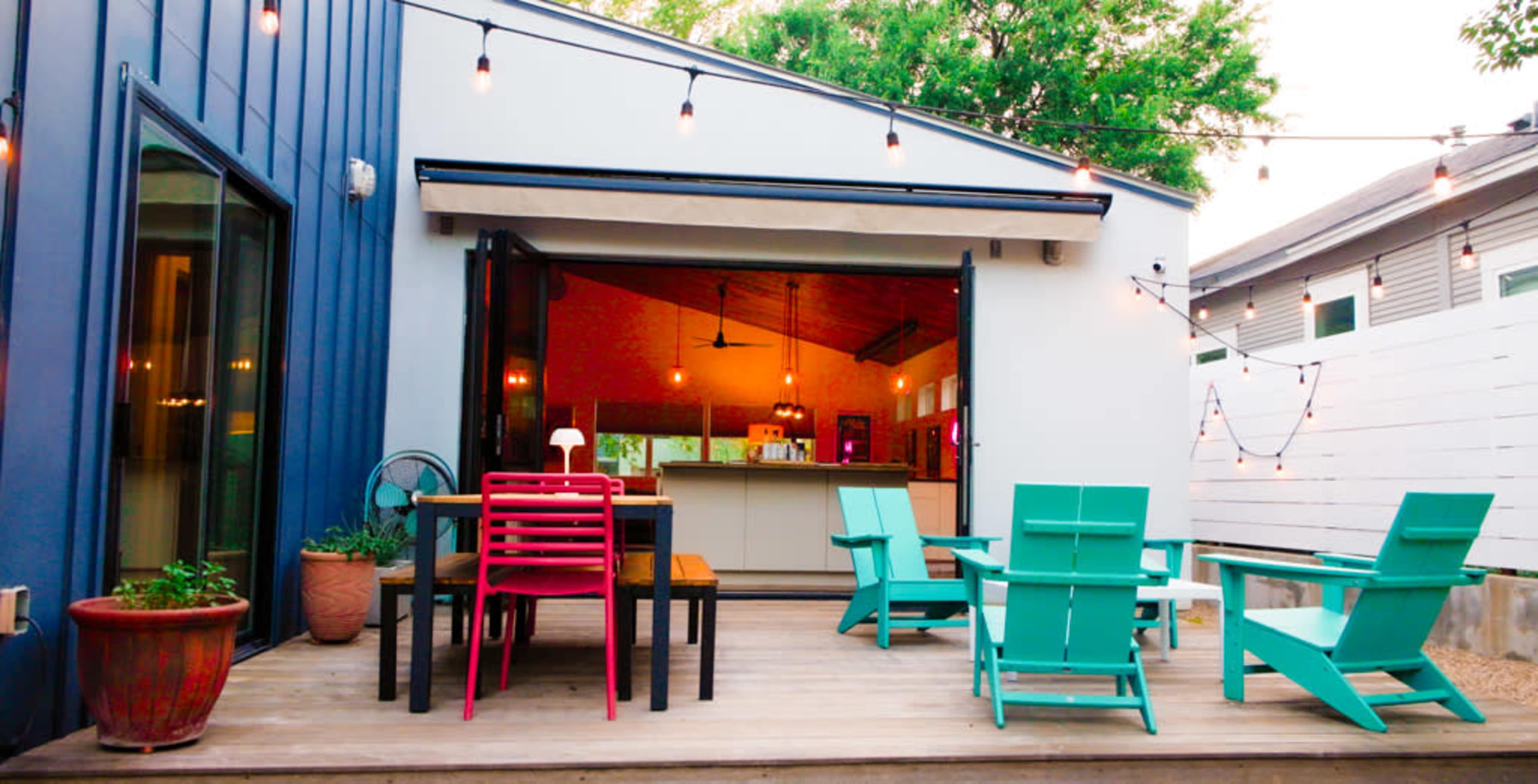 The image shows a backyard patio with colorful chairs and tables, surrounded by string lights and leading to an indoor kitchen area.
