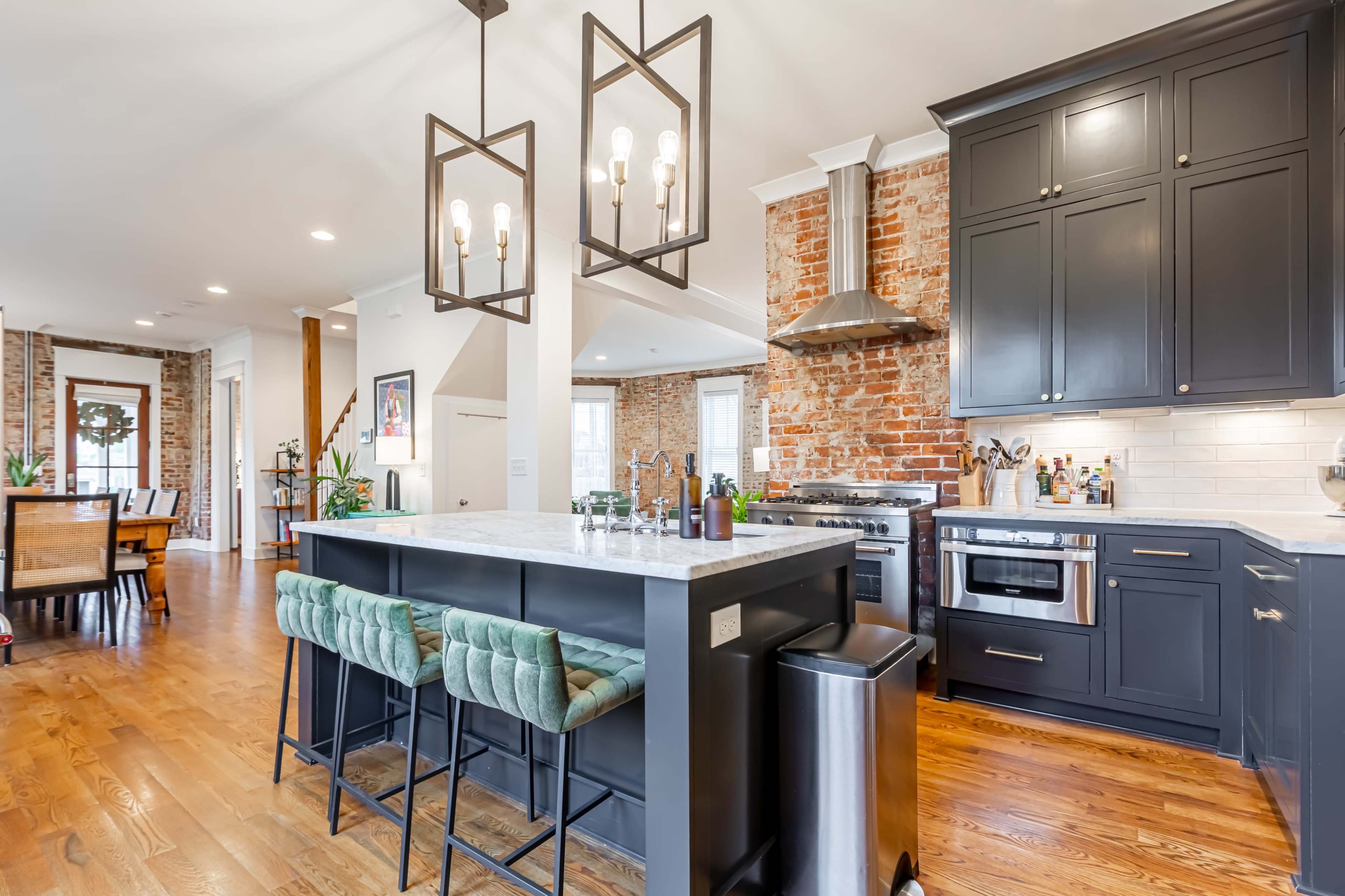 The image shows a modern kitchen featuring a large island with green bar stools, dark cabinetry, and exposed brick walls.