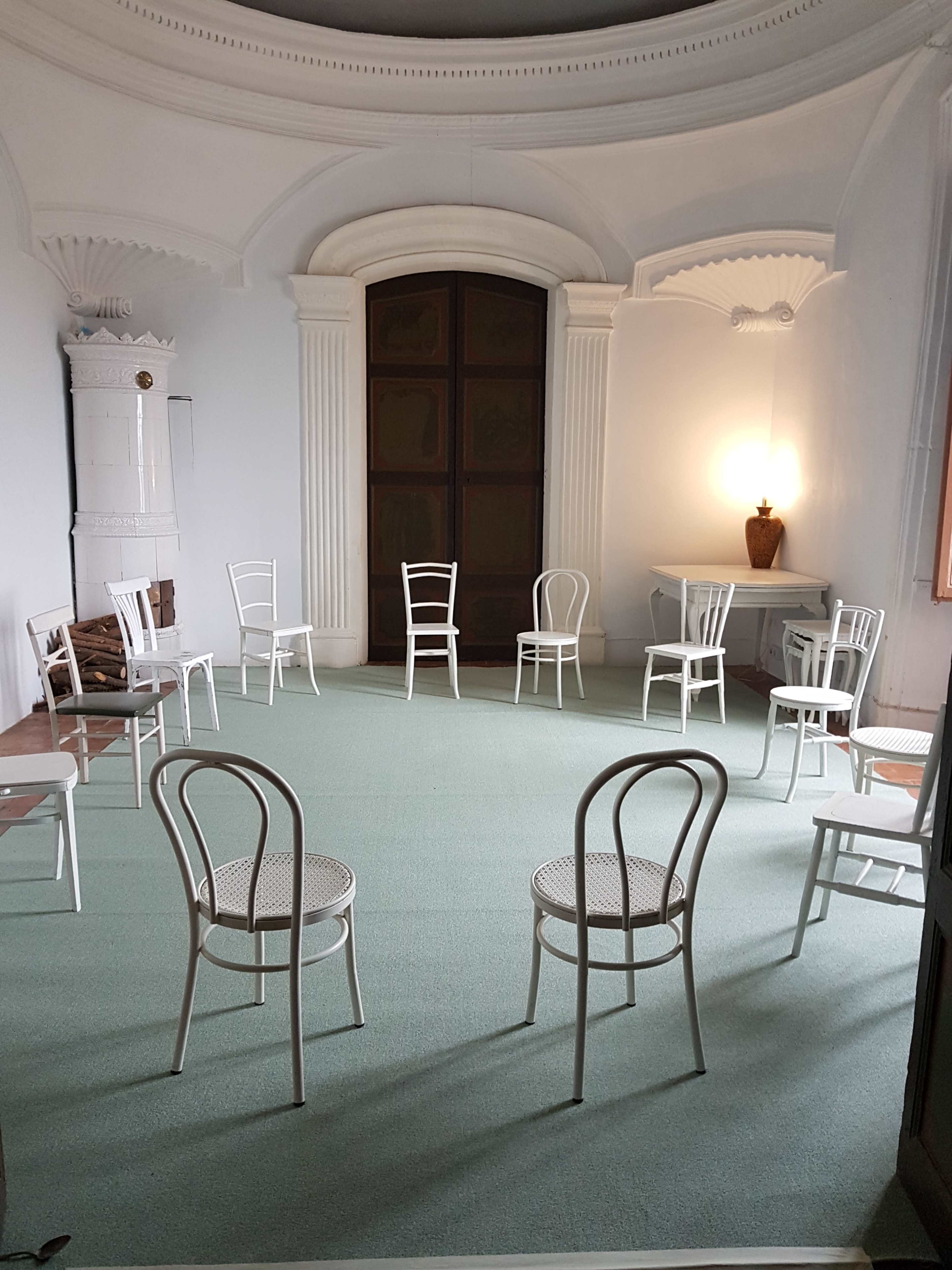 The room features a circular arrangement of white chairs on a green carpet, with a large wooden door and a lamp illuminating one corner.