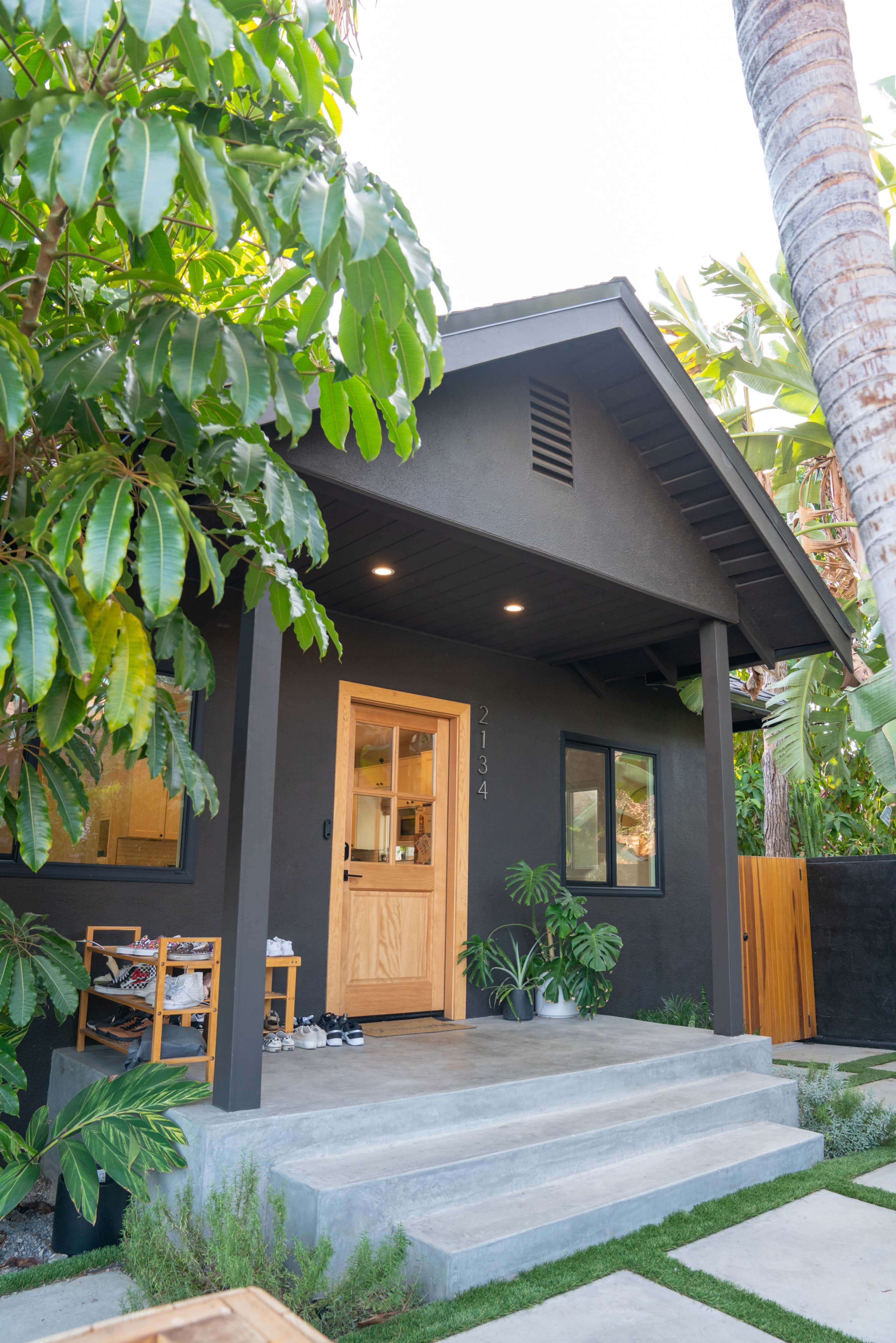 A modern black house with a wooden door and concrete steps is surrounded by lush greenery.