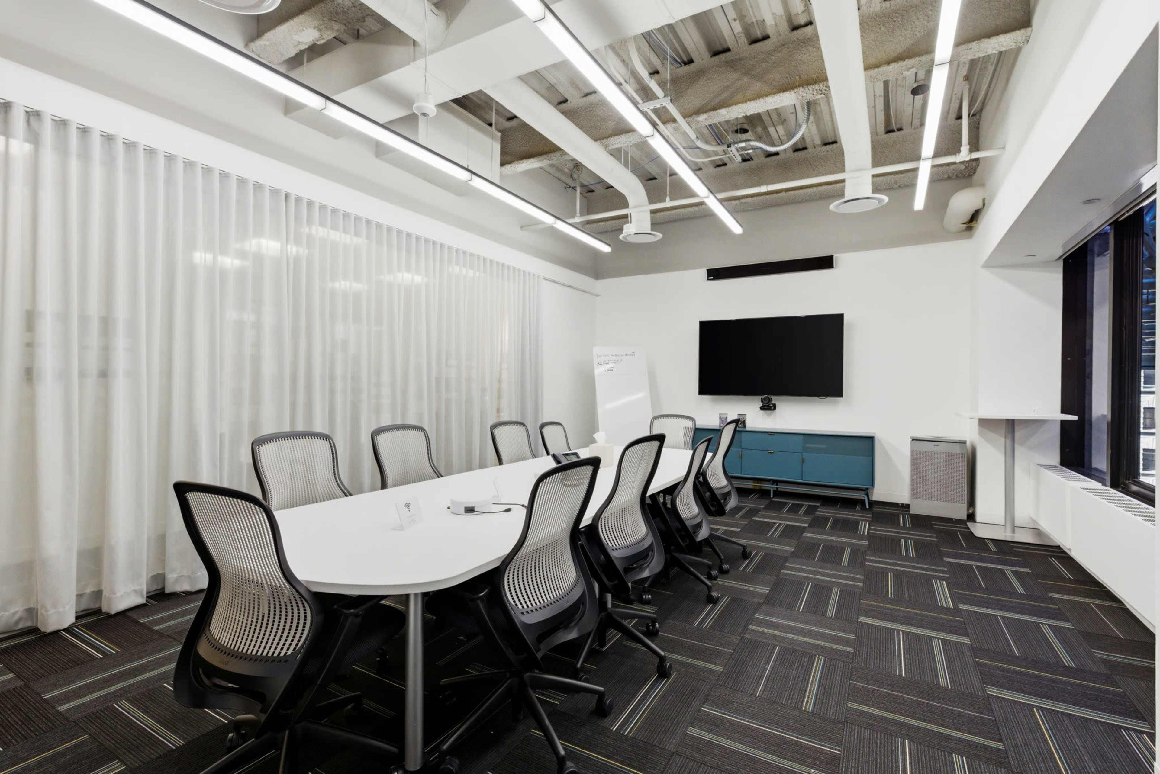 The image shows a modern conference room with a long table, ergonomic chairs, a large screen on the wall, and large windows letting in natural light.