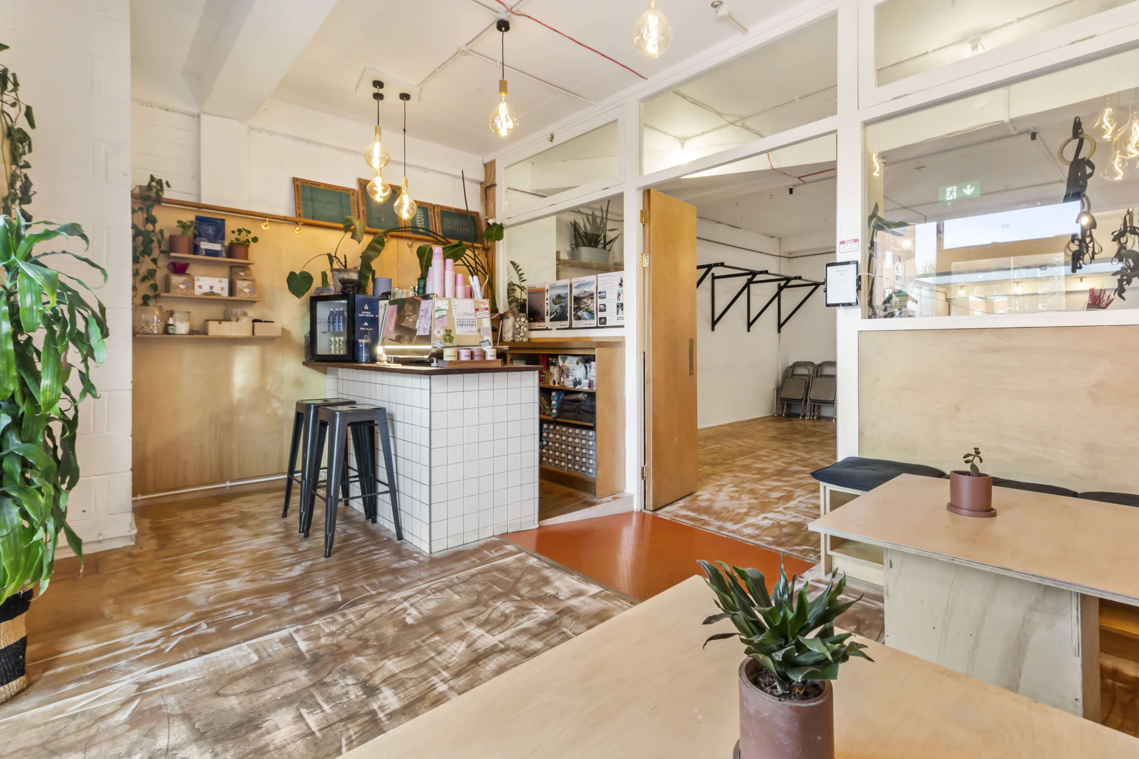 A modern café interior featuring wooden tables, bar stools, and plants, with a counter displaying various items and seating areas in the background.