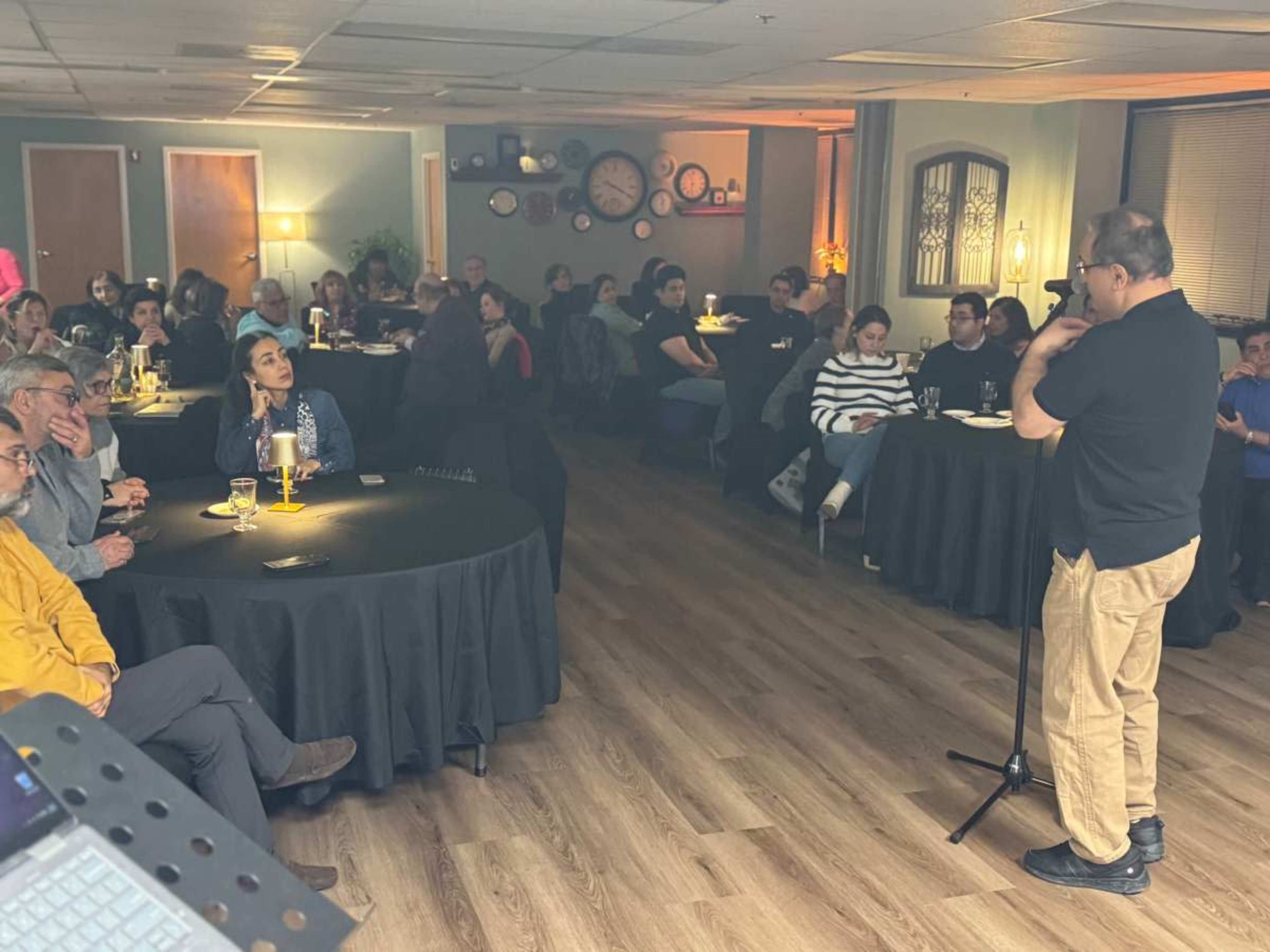 A diverse audience sits at tables in a dimly lit room while a speaker stands on stage, engaging with the crowd.