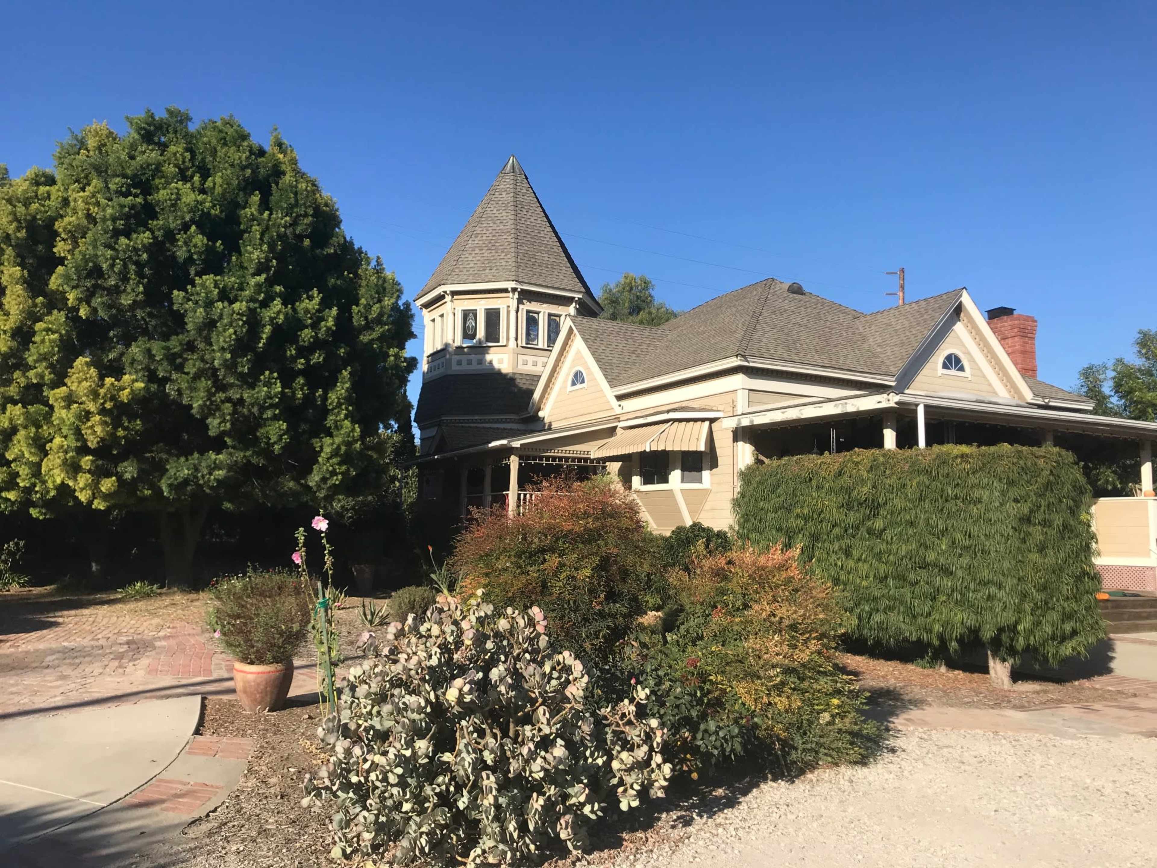 A Victorian-style house with a peaked tower is surrounded by well-maintained shrubs and trees.