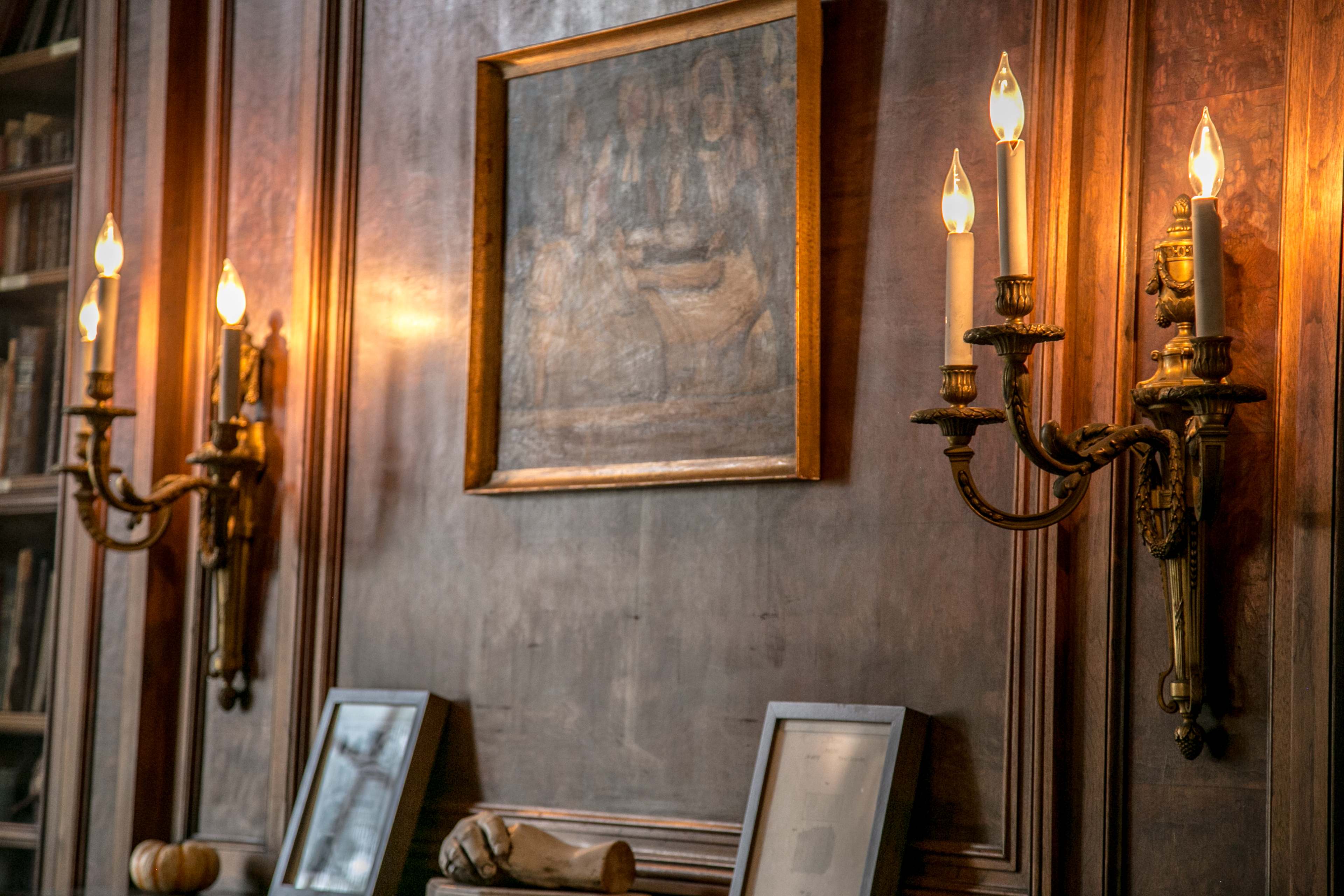 A wooden wall adorned with a painting and two antique sconces illuminating the space.