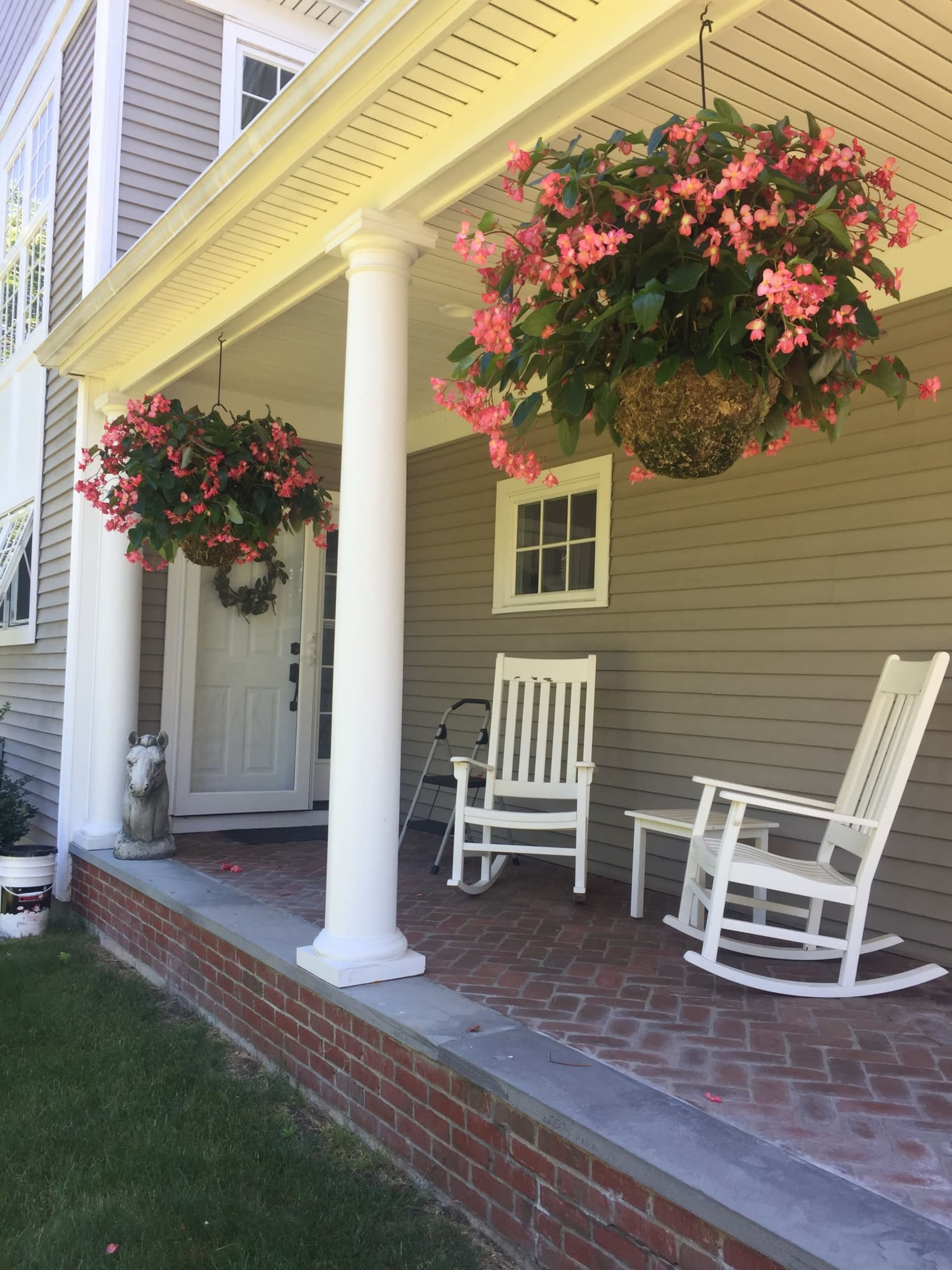 A front porch with two white rocking chairs, hanging floral baskets, and a decorative statue beside the entrance.