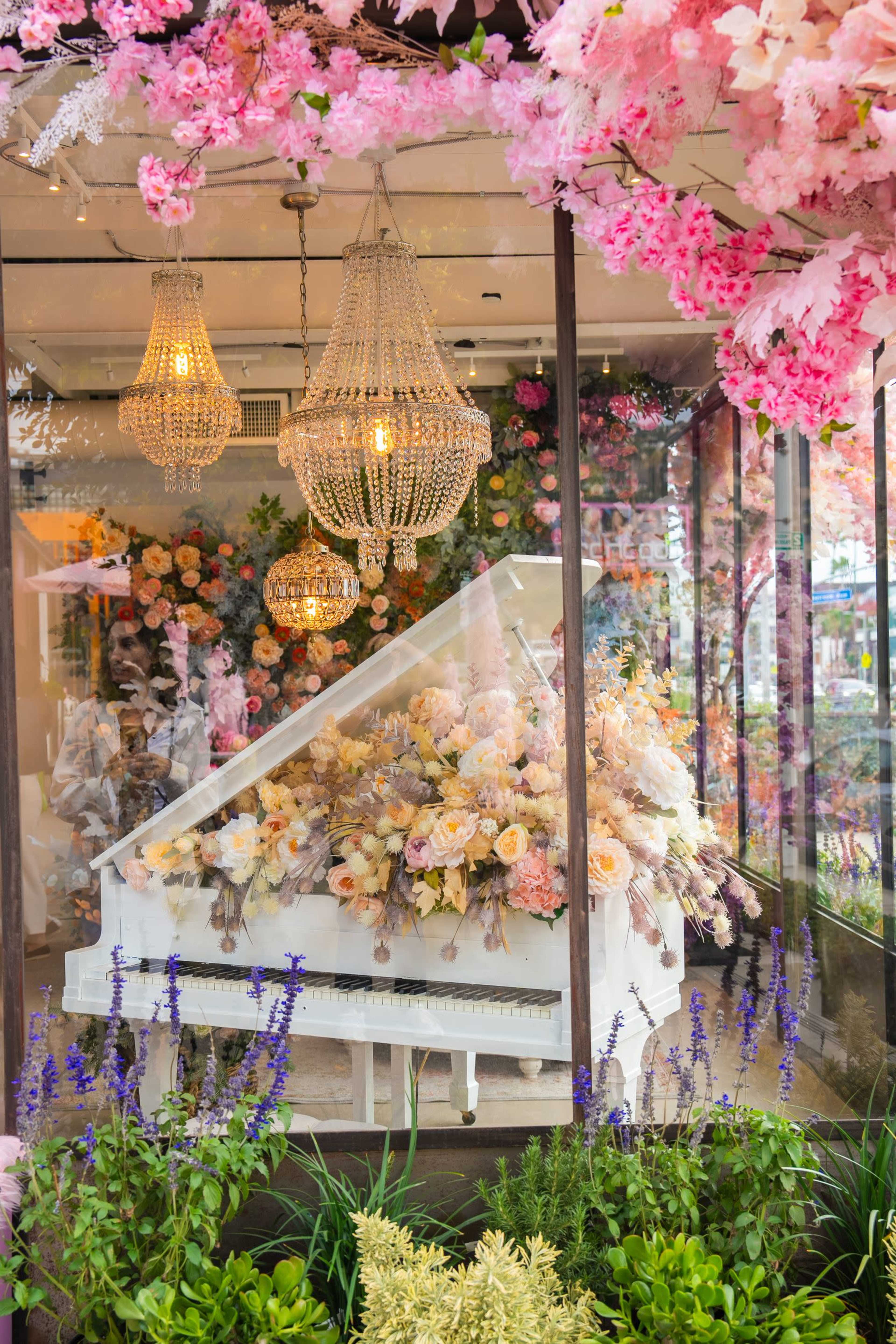 A white grand piano adorned with flowers is placed in a glass display surrounded by chandeliers and floral decorations.