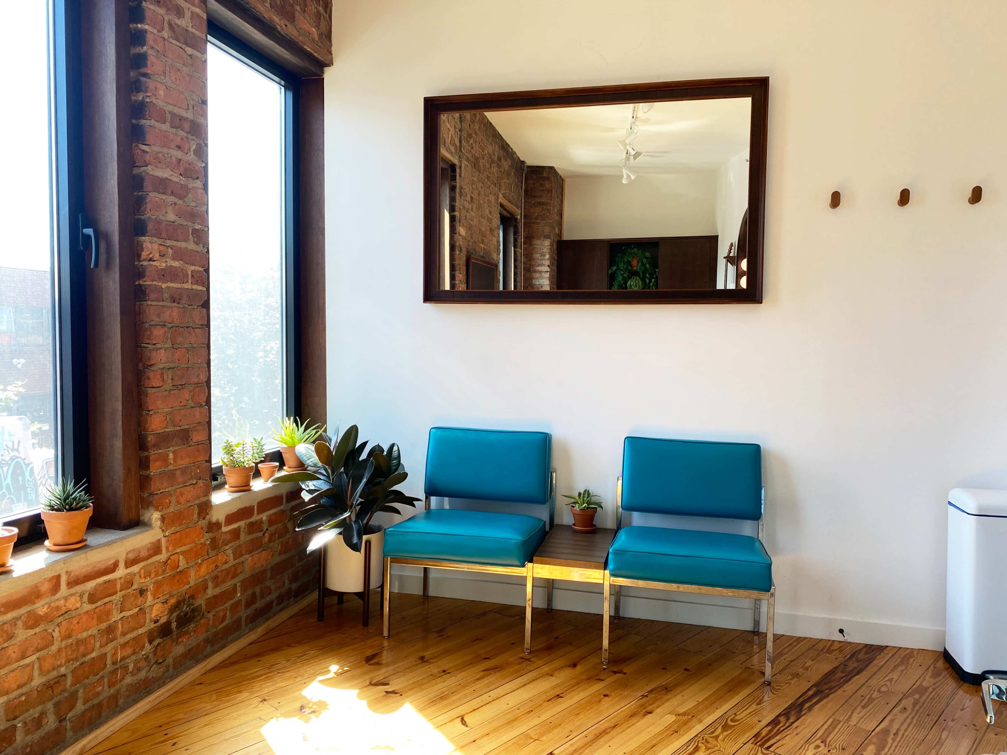 Two blue chairs positioned against a wall with a large mirror above them, surrounded by potted plants on the windowsill and a brick backdrop.