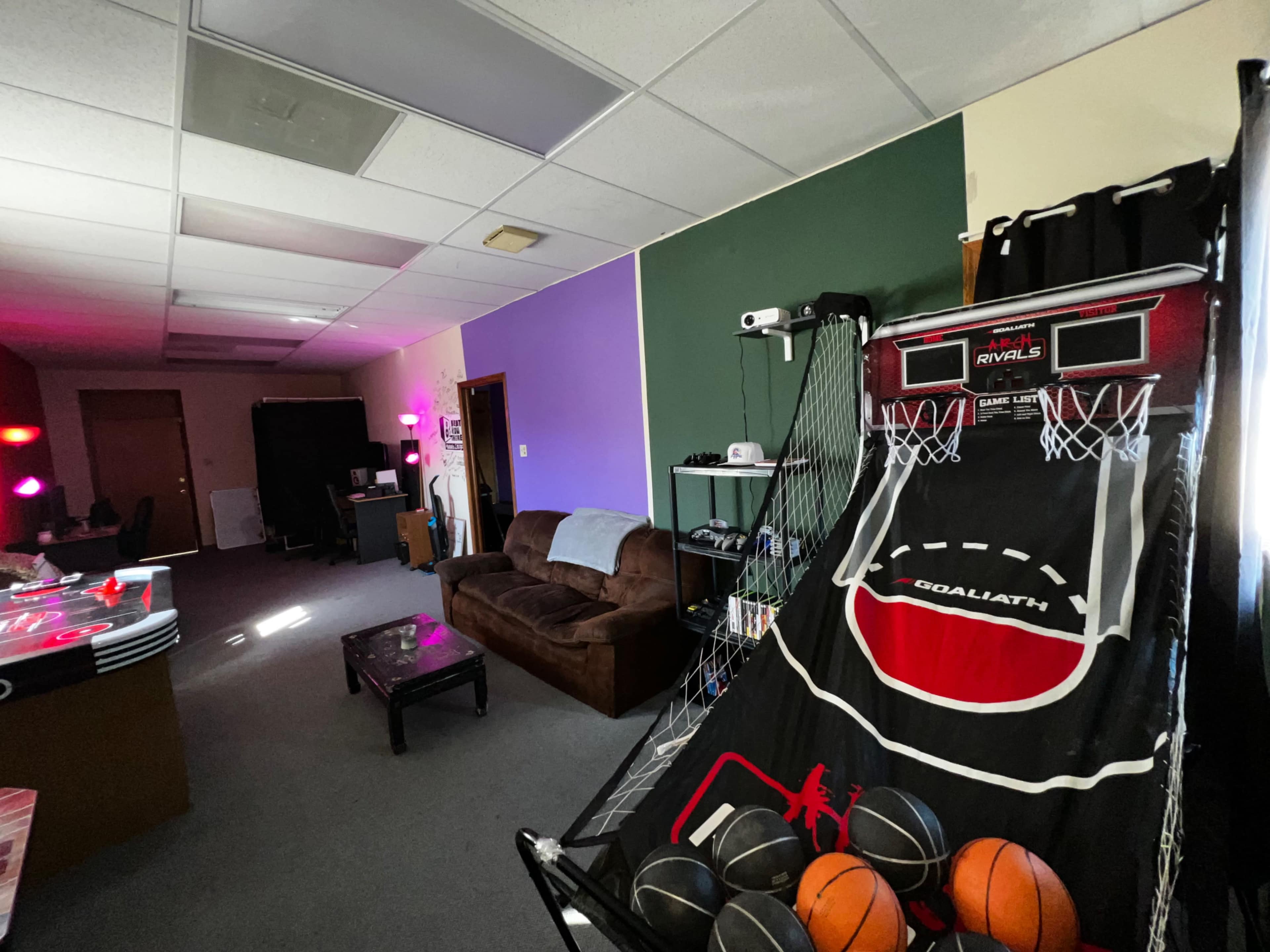 A room featuring a basketball arcade game, a brown couch, and purple-painted walls, with various items set up in the background.