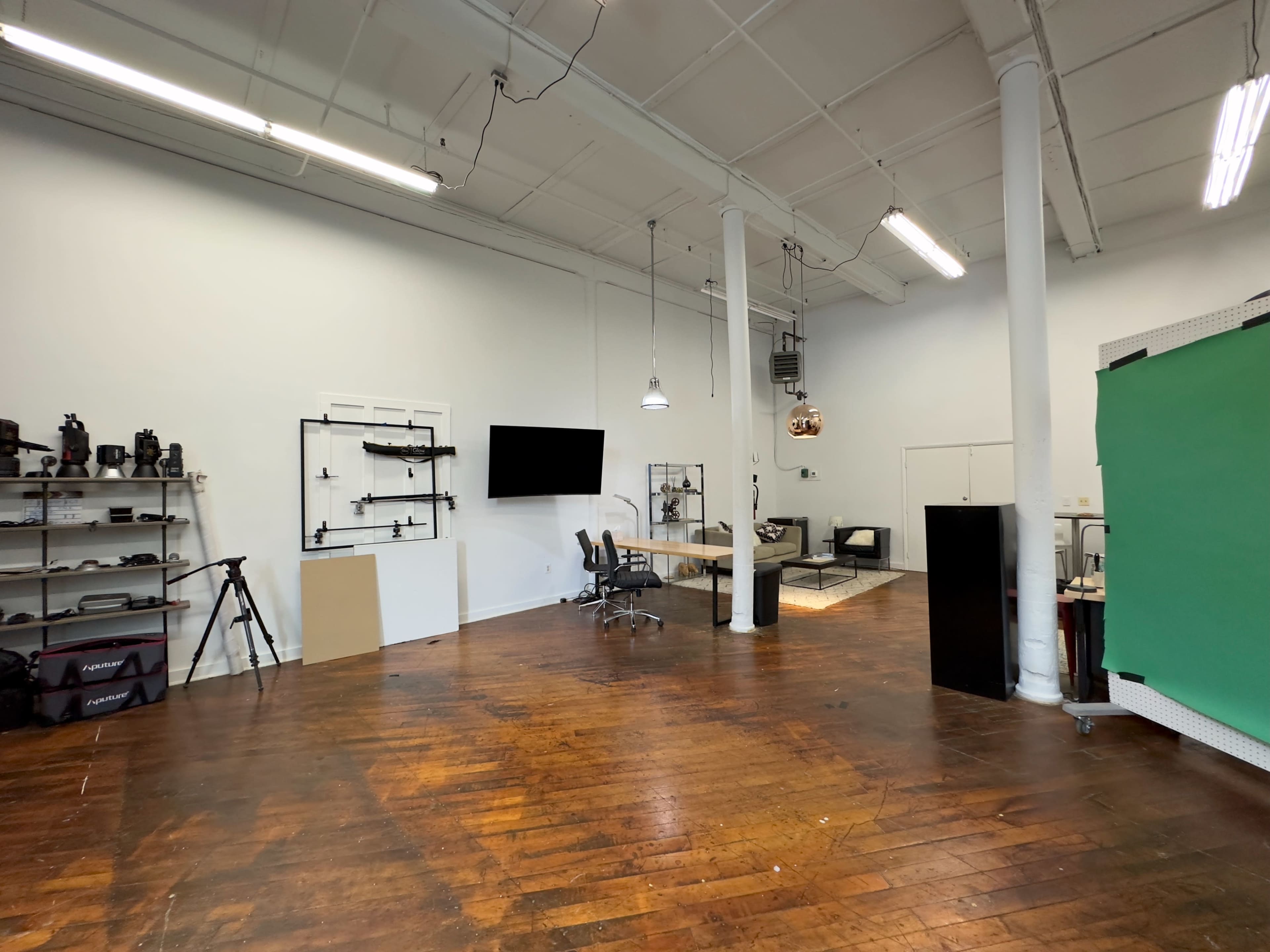 A spacious, well-lit studio with wooden floors, a green backdrop, a large screen, a desk with chairs, and various equipment arranged on shelves.