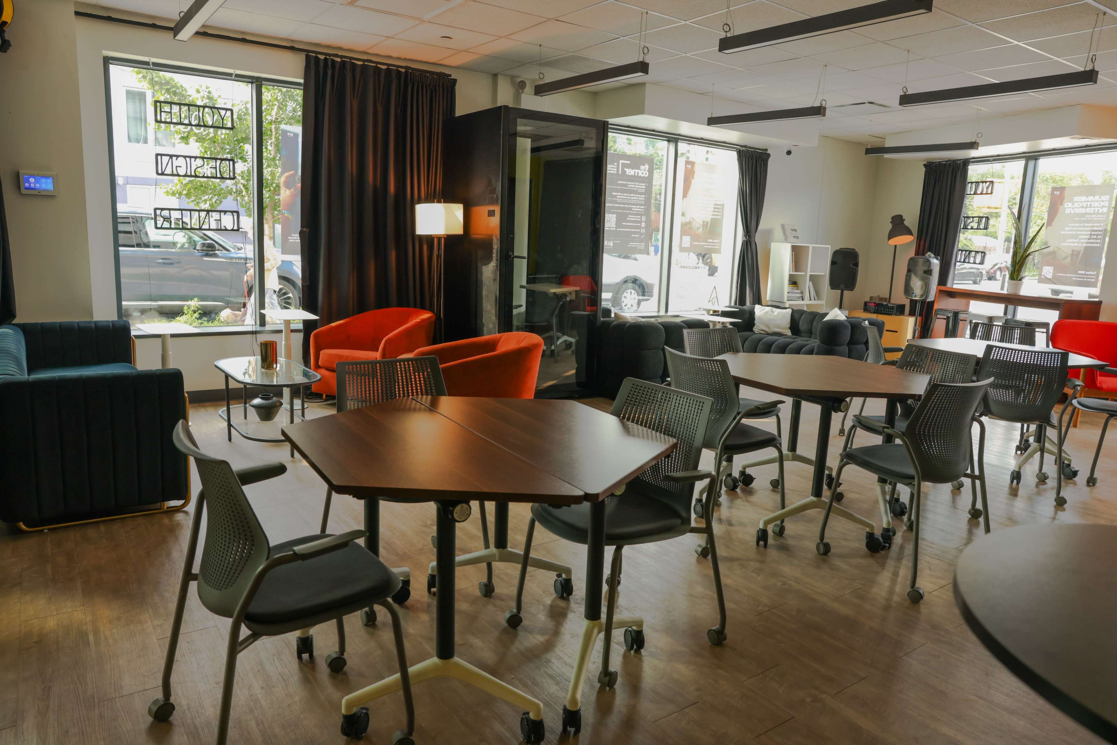 The image shows a modern co-working space with various seating arrangements, including colorful chairs and tables, illuminated by natural light from large windows.