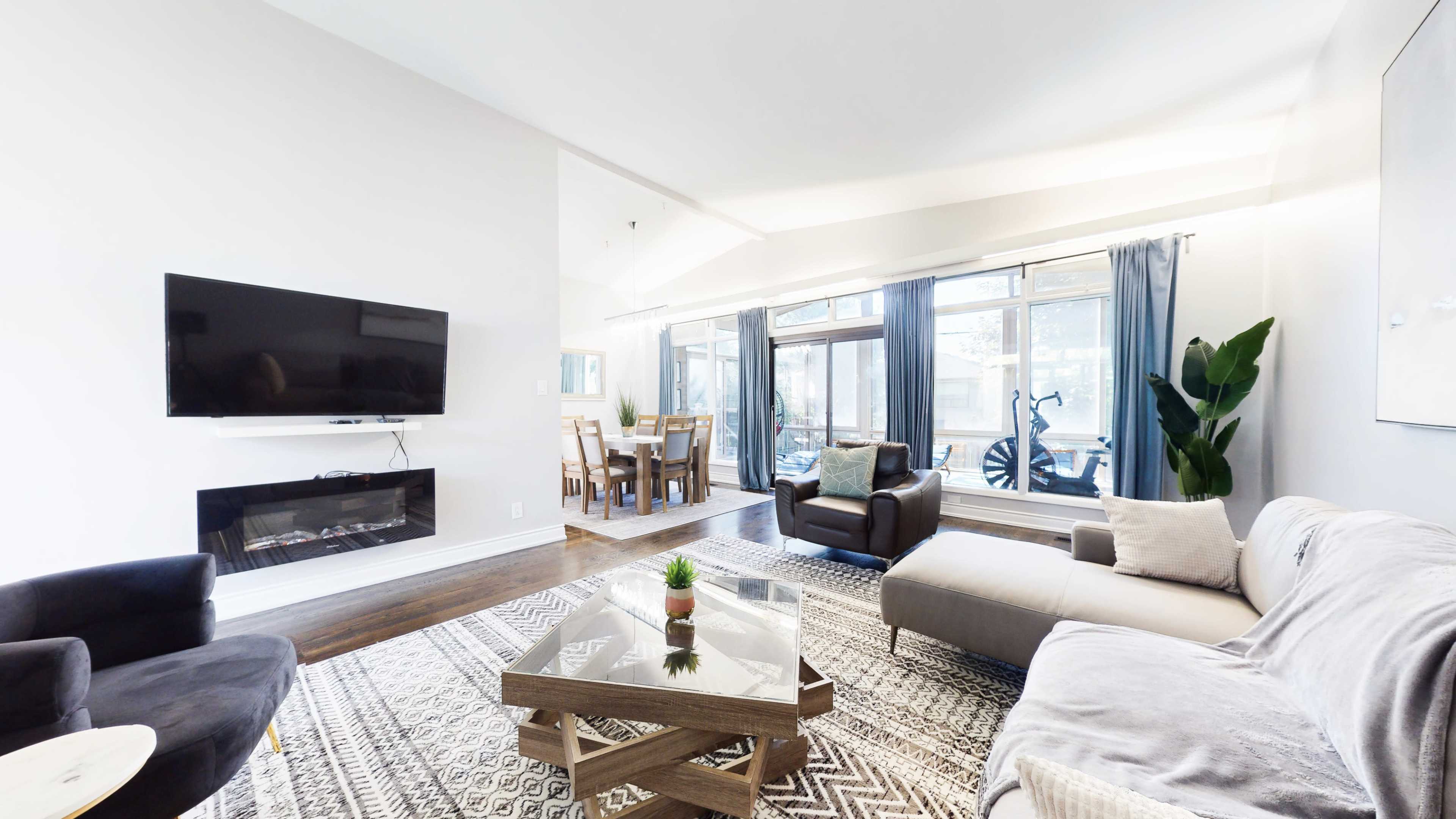 The image shows a modern living room with a sectional sofa, a glass coffee table, and a large wall-mounted television, alongside a dining area with a visible table and chairs.