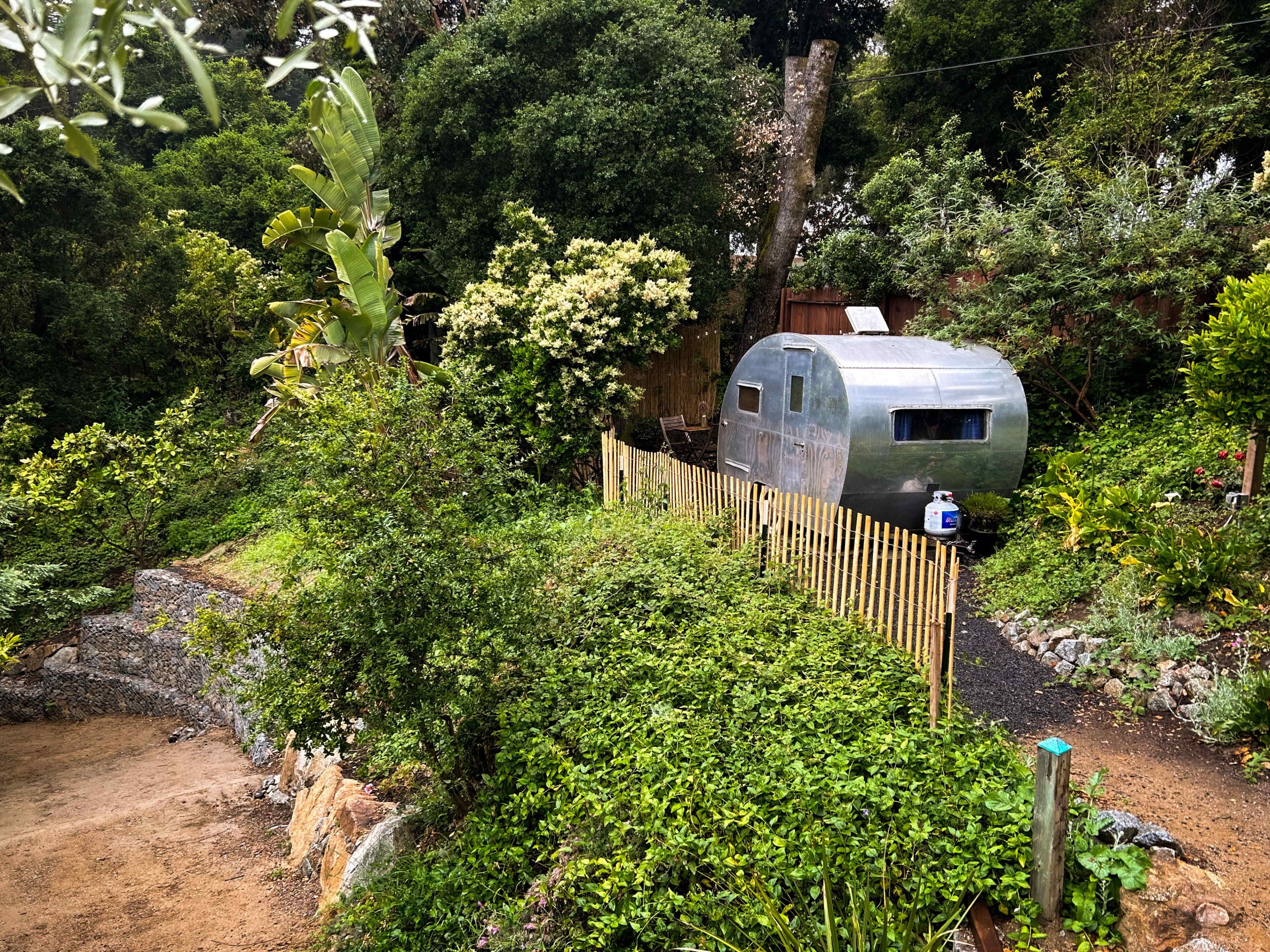 A vintage silver trailer is positioned near a wooden fence amidst lush greenery and trees.