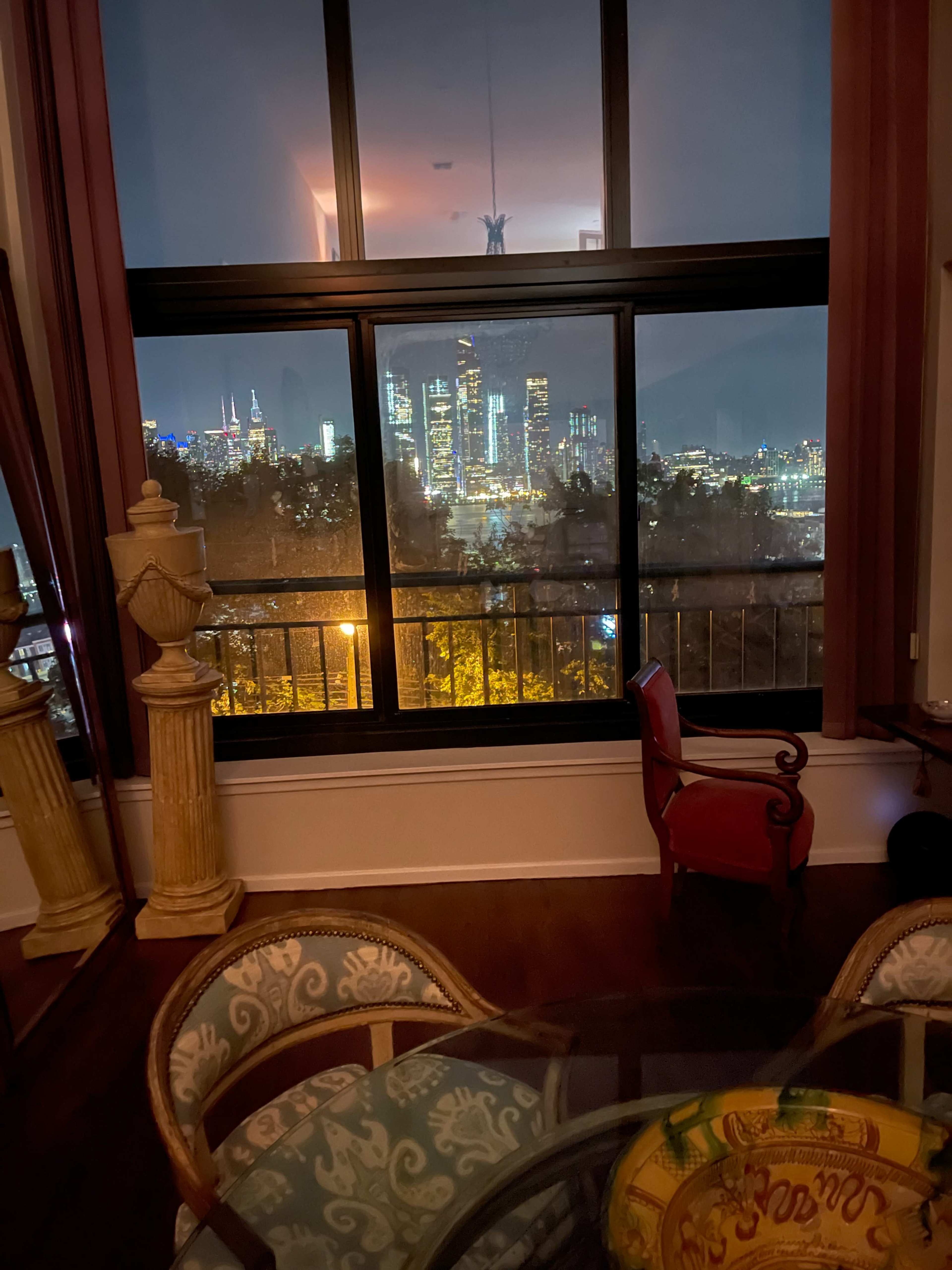 The image shows a view from an interior space with a dining table and a chair, overlooking a city skyline at night through a large window.