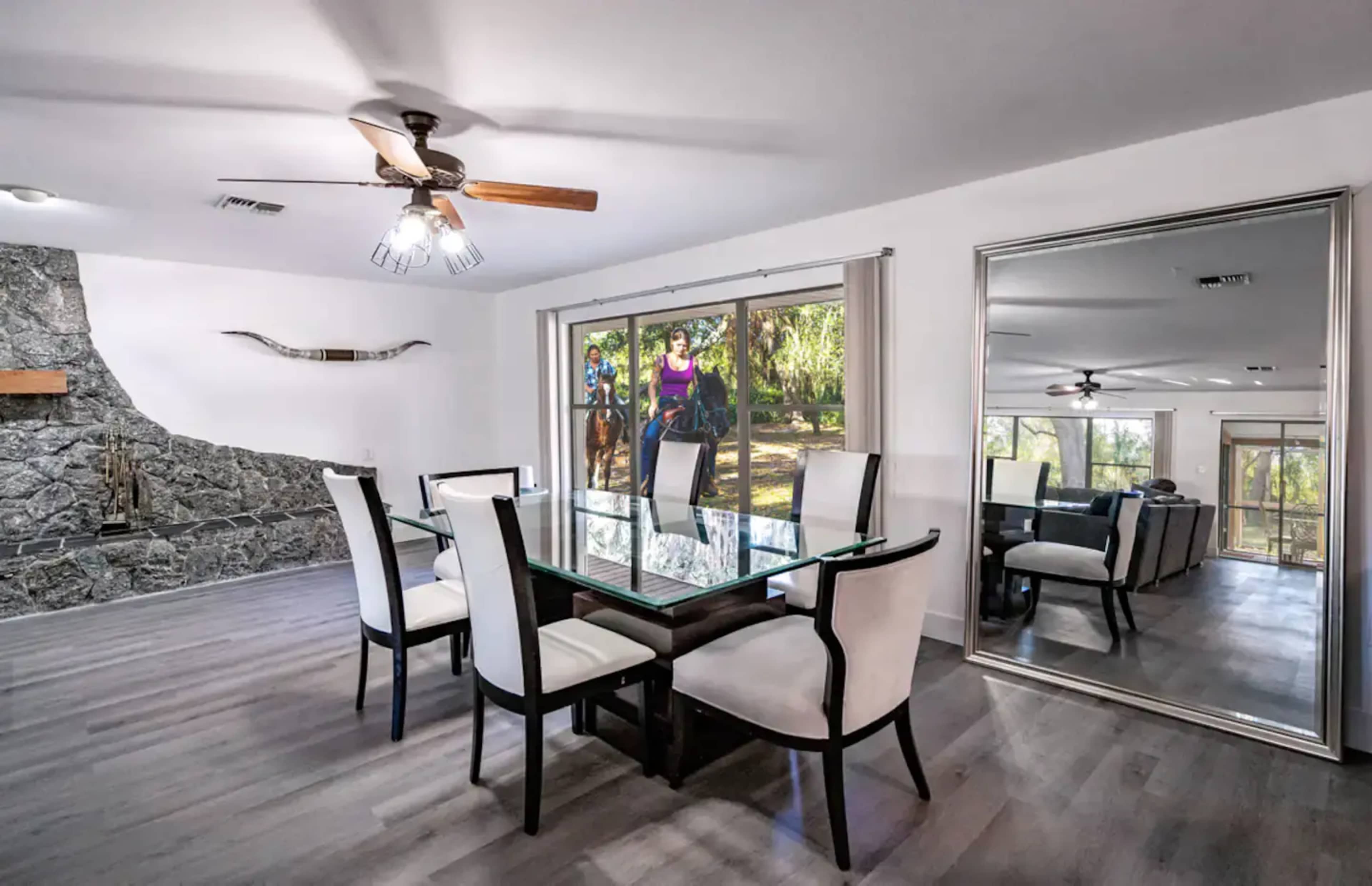 A dining area features a glass table surrounded by six chairs, with a view of two riders on horseback visible through large windows.
