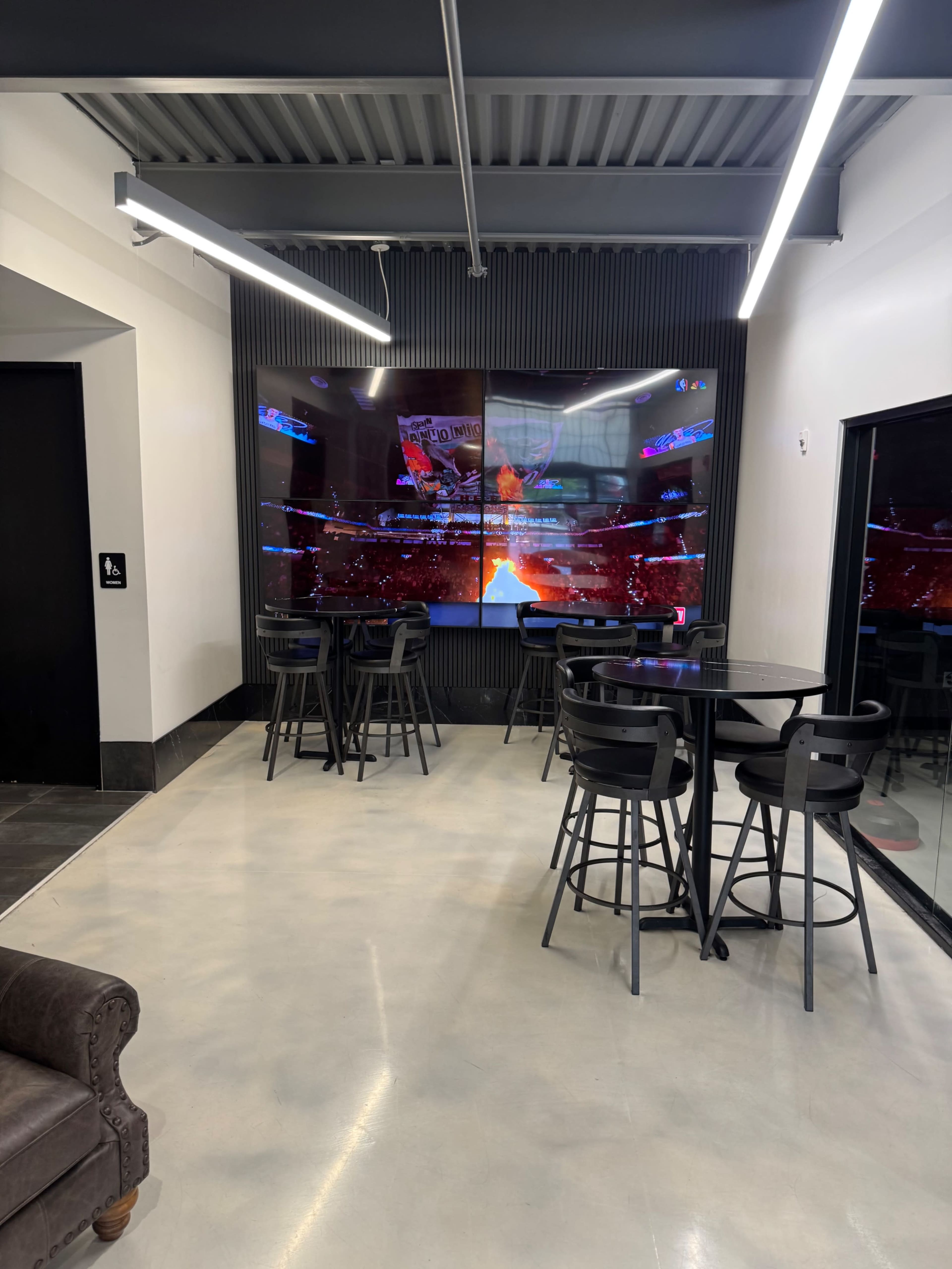 The image shows a modern lounge area featuring black bar stools and tables in front of a large, multi-screen display showing a sports event.