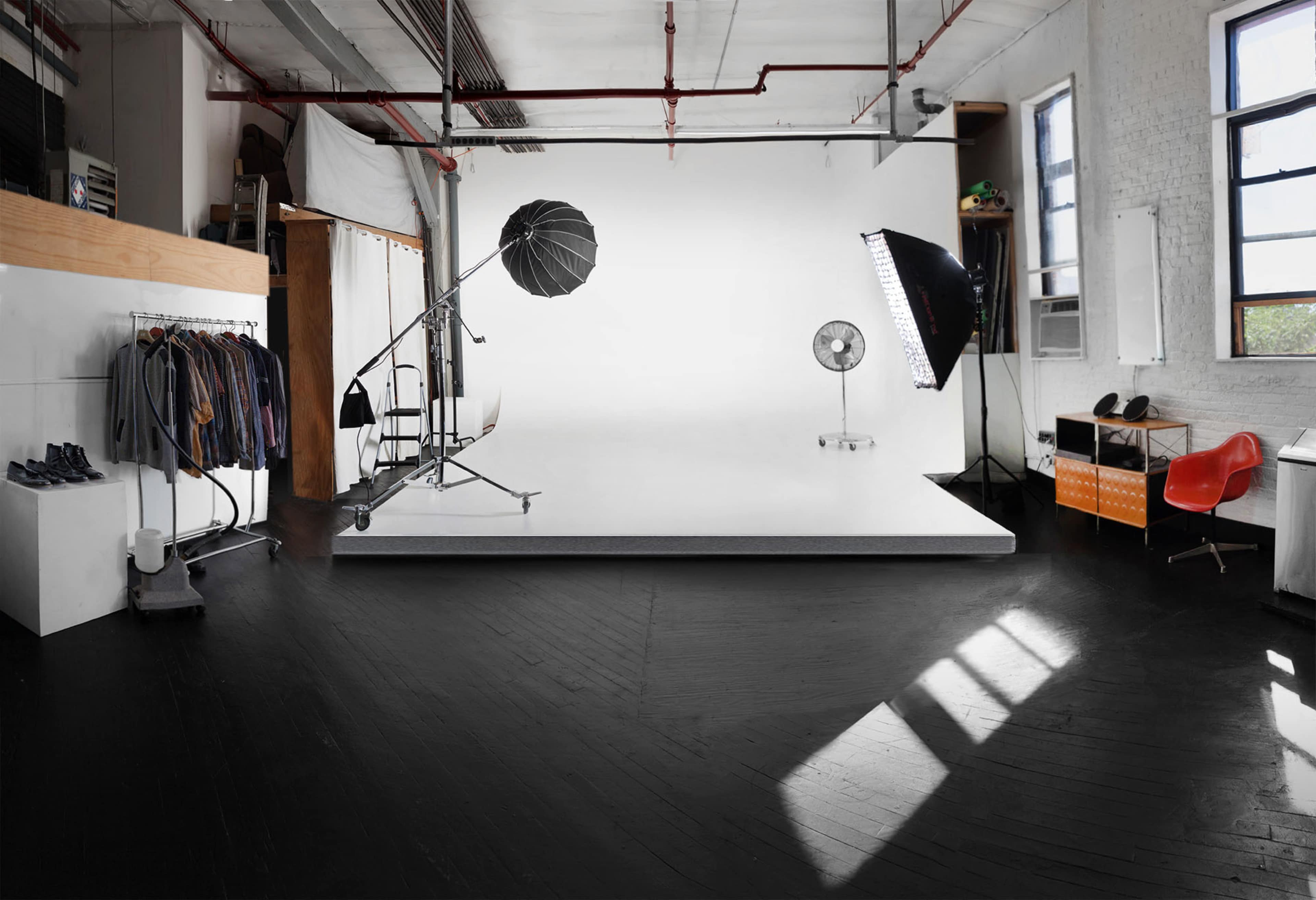A photography studio features a large white backdrop, various clothing on hangers, lighting equipment, and a fan positioned on a wooden platform.