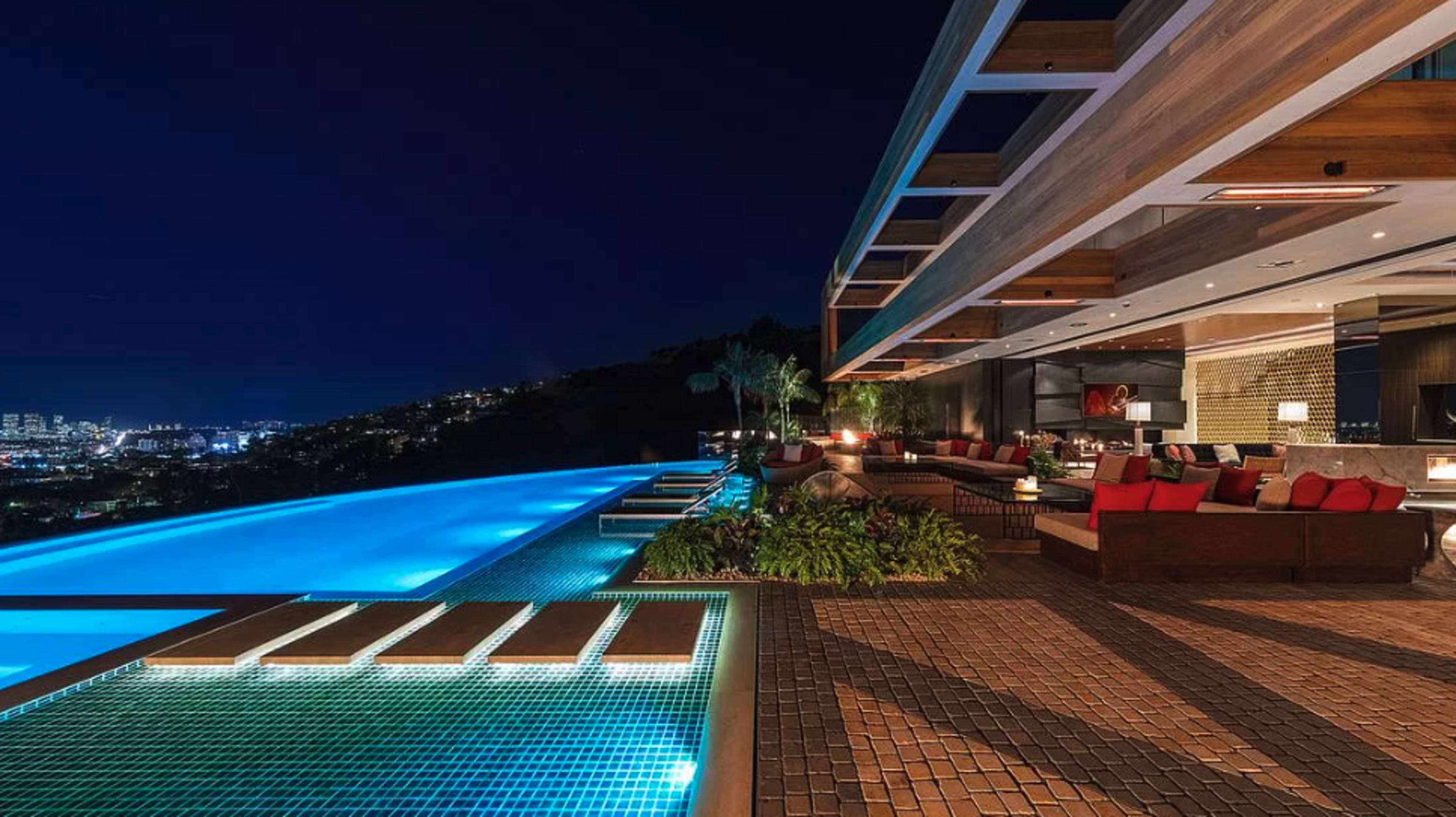 The image shows a modern outdoor pool area at night, featuring an infinity pool with illuminated water, adjacent lounge seating, and a spacious patio with a view of a city skyline.