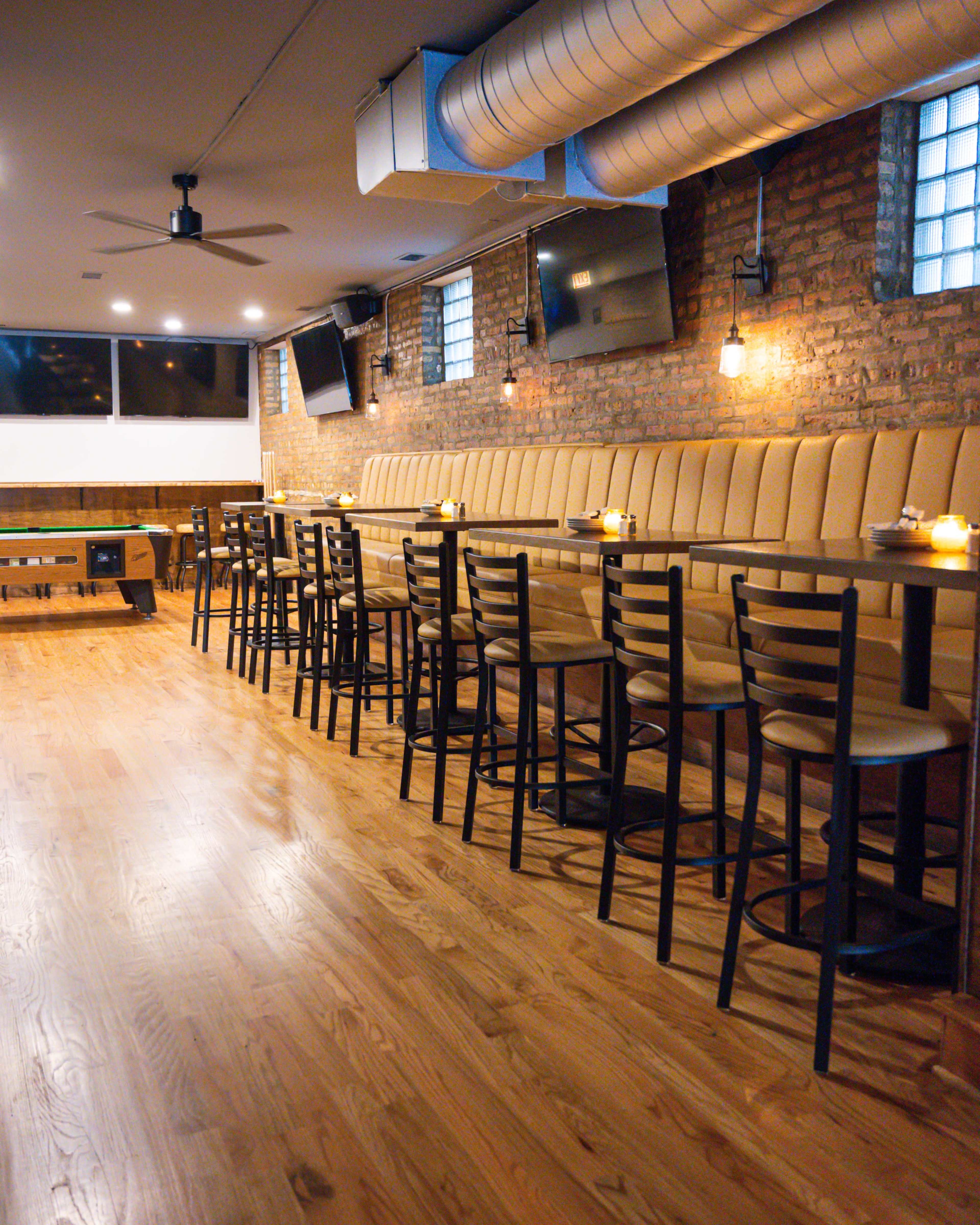The image shows a modern bar interior with a long padded seating area, high tables, and wooden floors, illuminated by pendant lighting.