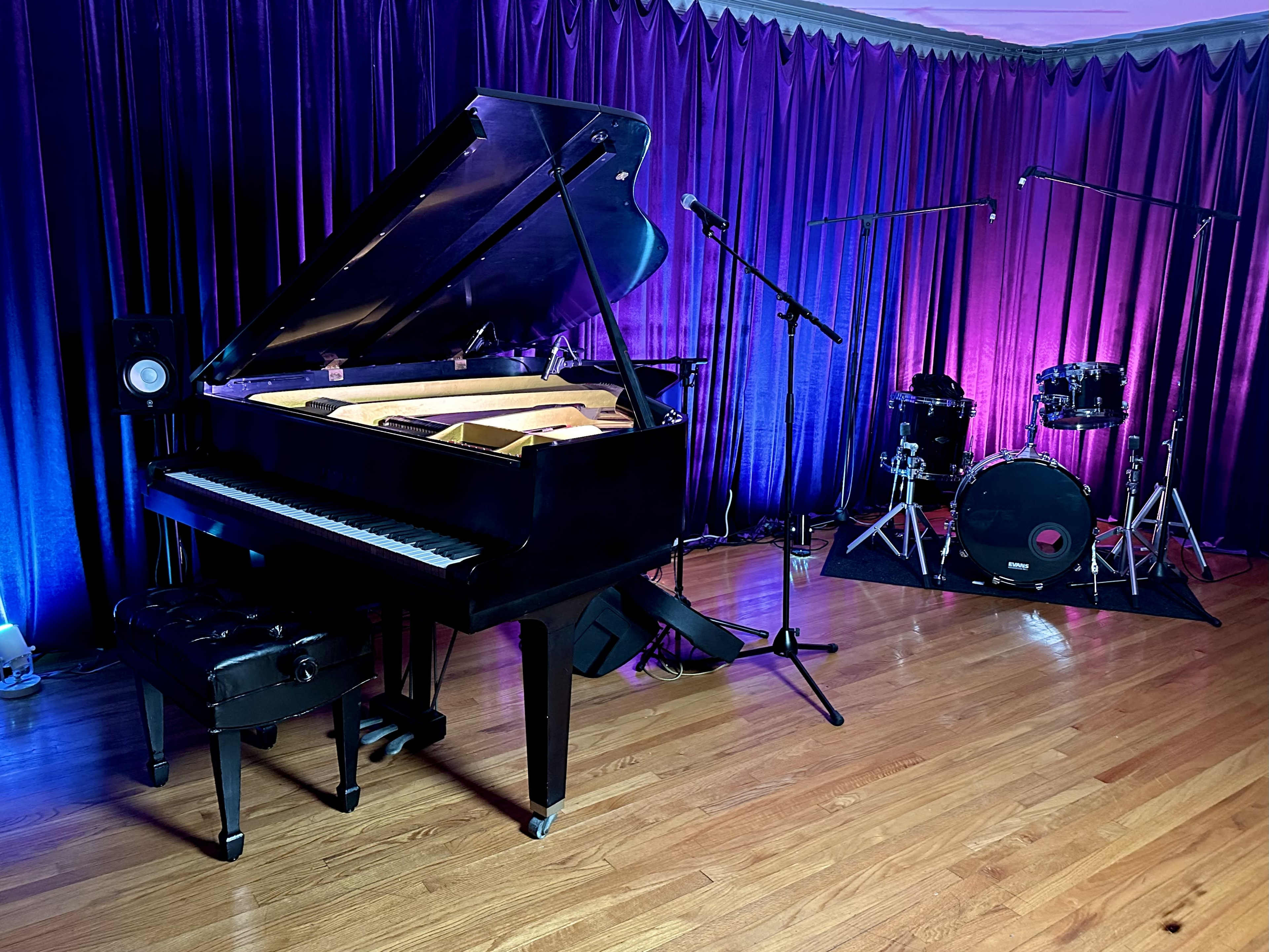 A black grand piano sits on polished wooden flooring in a room with purple curtains and stage lighting, accompanied by microphone stands and a drum set in the background.