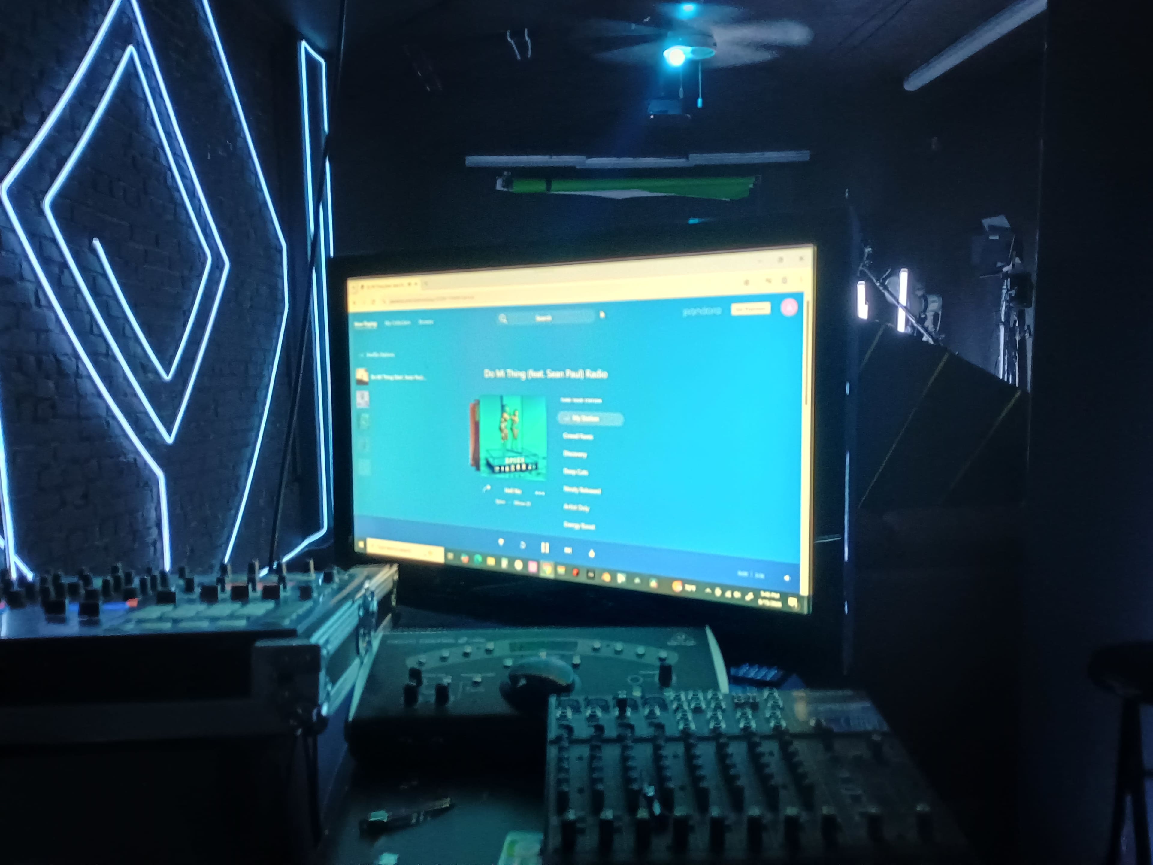 A large monitor displaying a music streaming service is positioned on a tabletop with audio equipment in a dimly lit room.