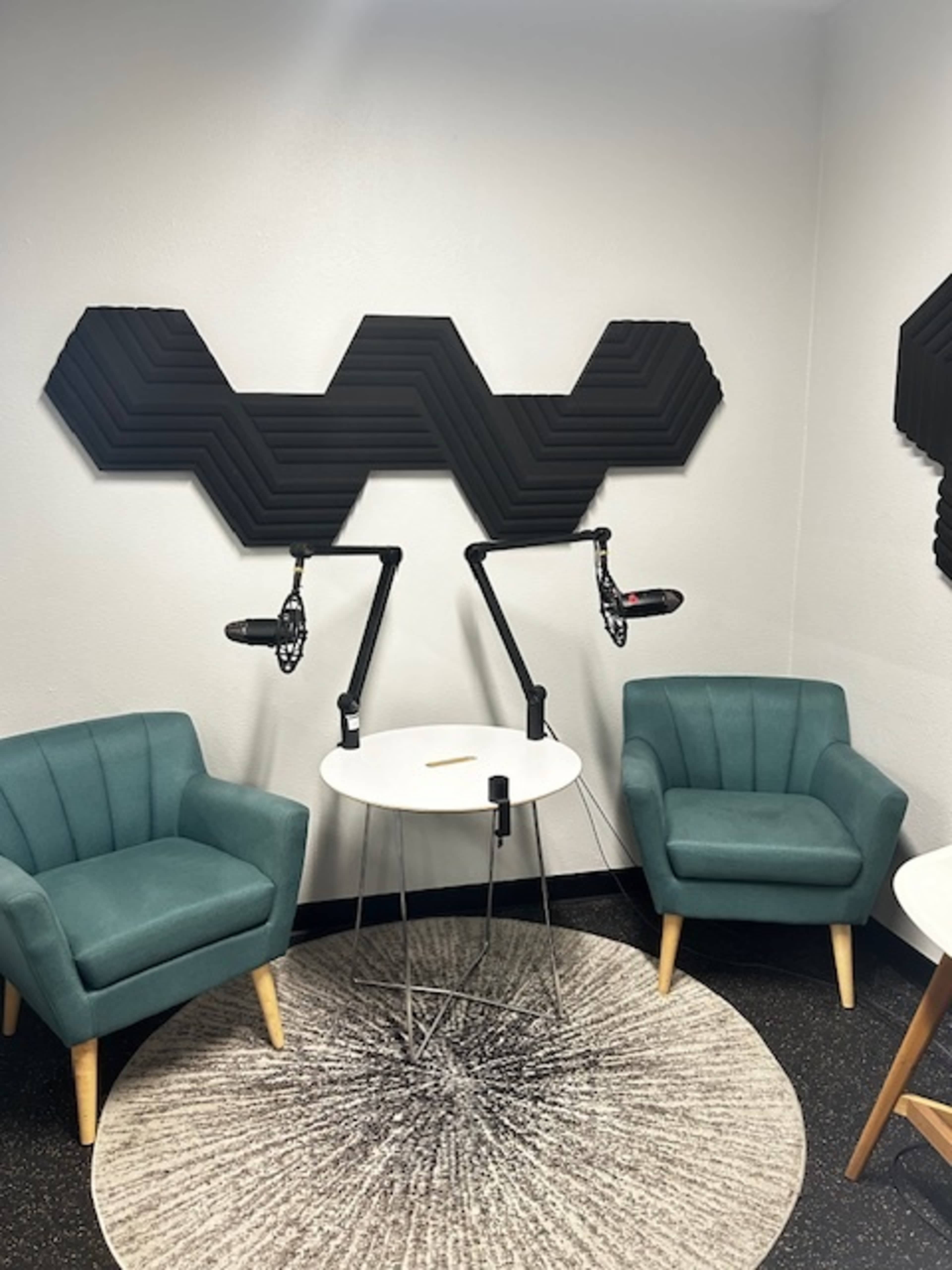 A small recording studio featuring two teal chairs, a round table, and soundproofing panels on the wall above microphone stands.