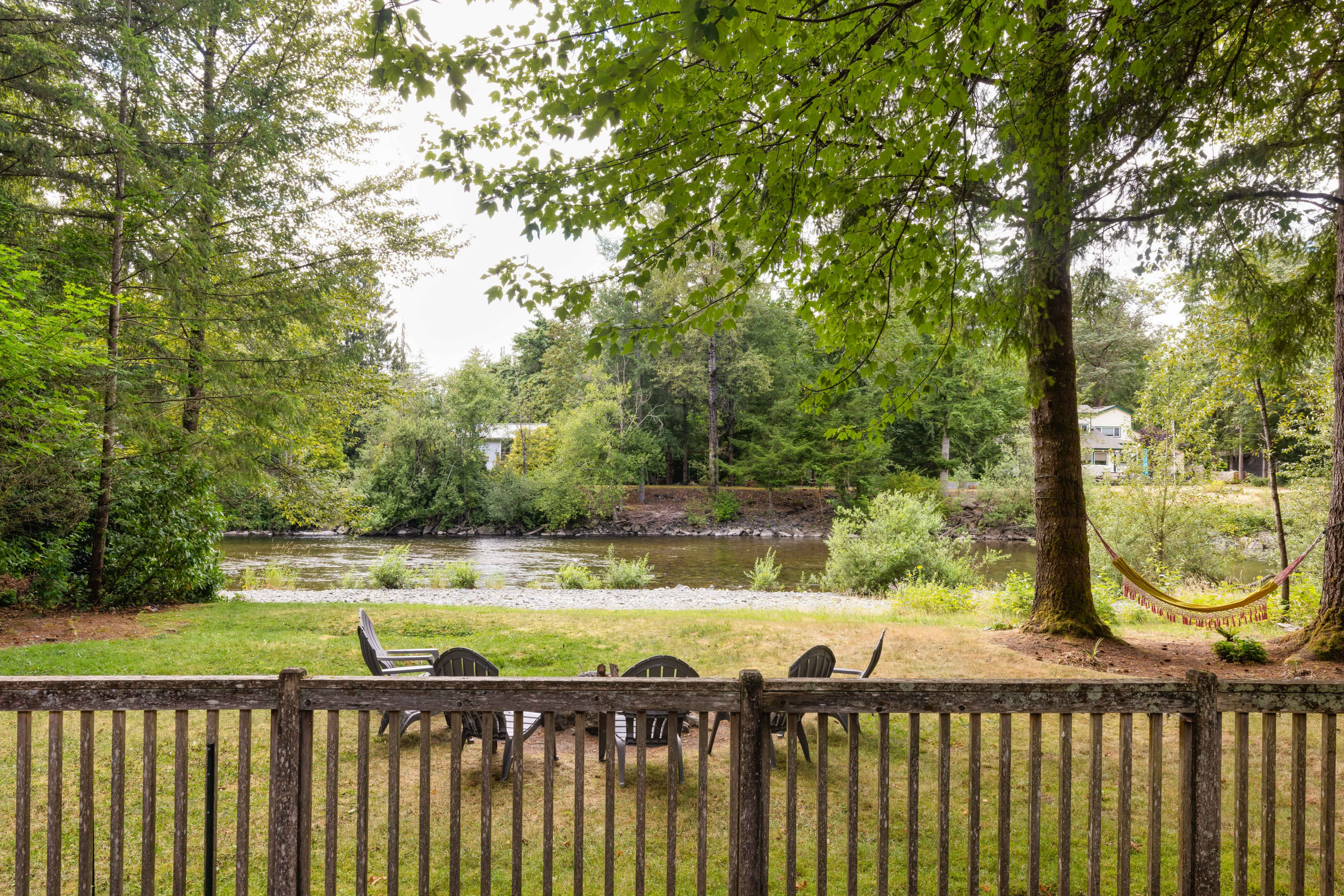 A fenced yard features a circular arrangement of chairs facing a river, surrounded by trees and a hammock nearby.