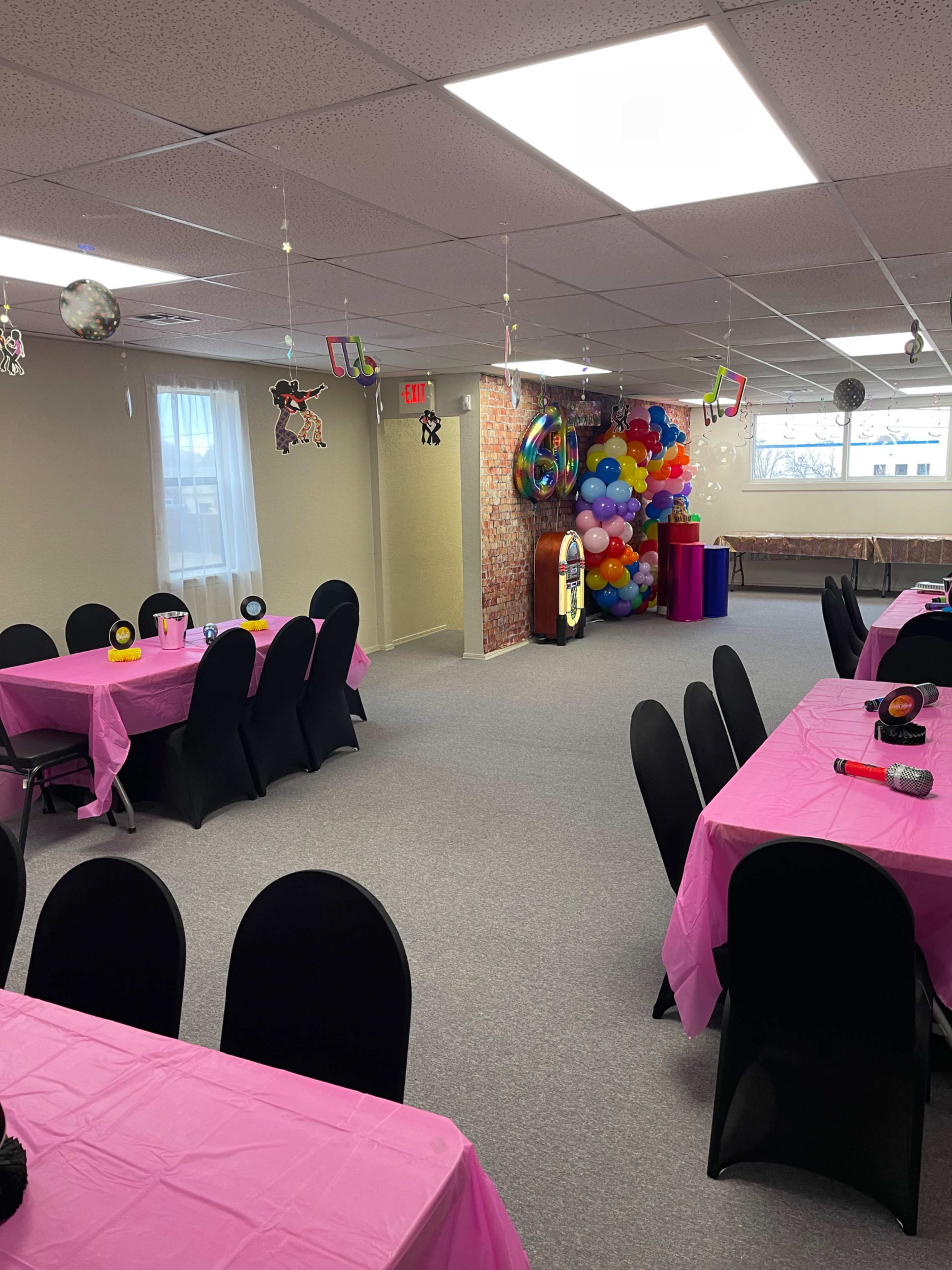 The image shows a spacious event room with pink tablecloths, chairs, and colorful decorations, including a large balloon arrangement.