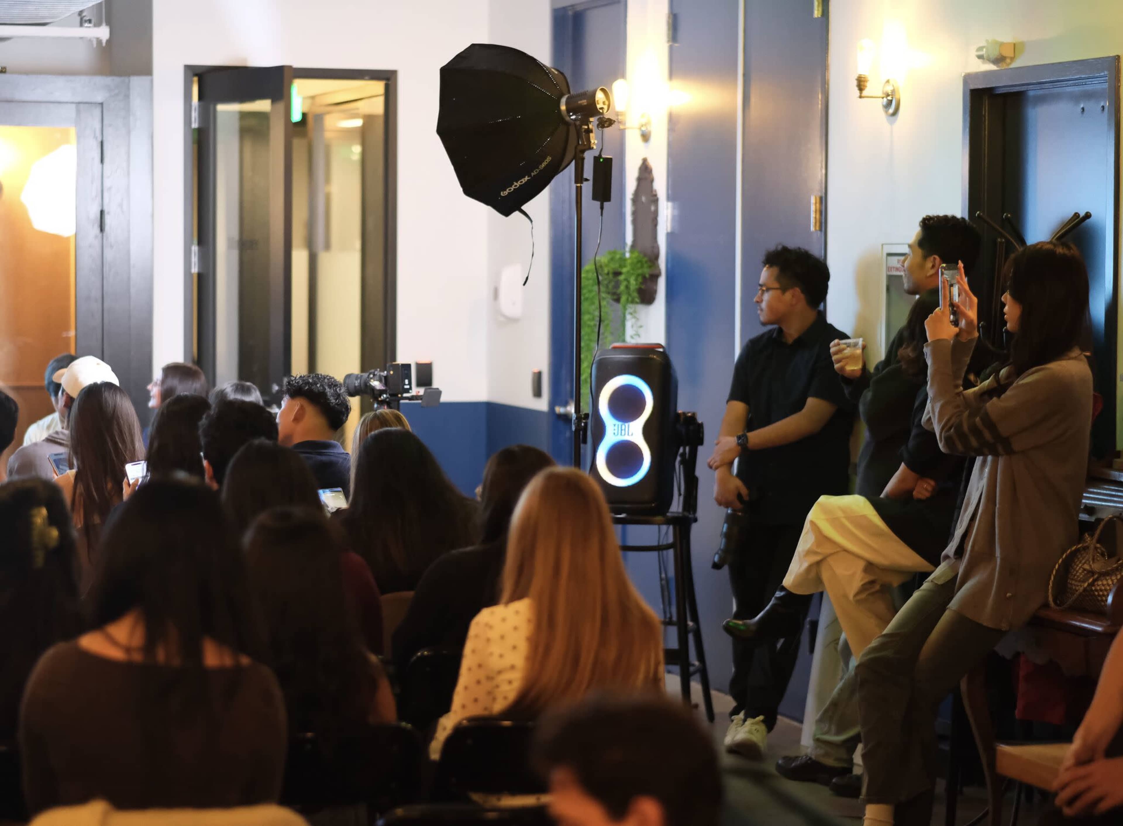 A crowd of people is seated in a room, some using their phones, while a few individuals stand near a photography setup with a softbox and a light source.
