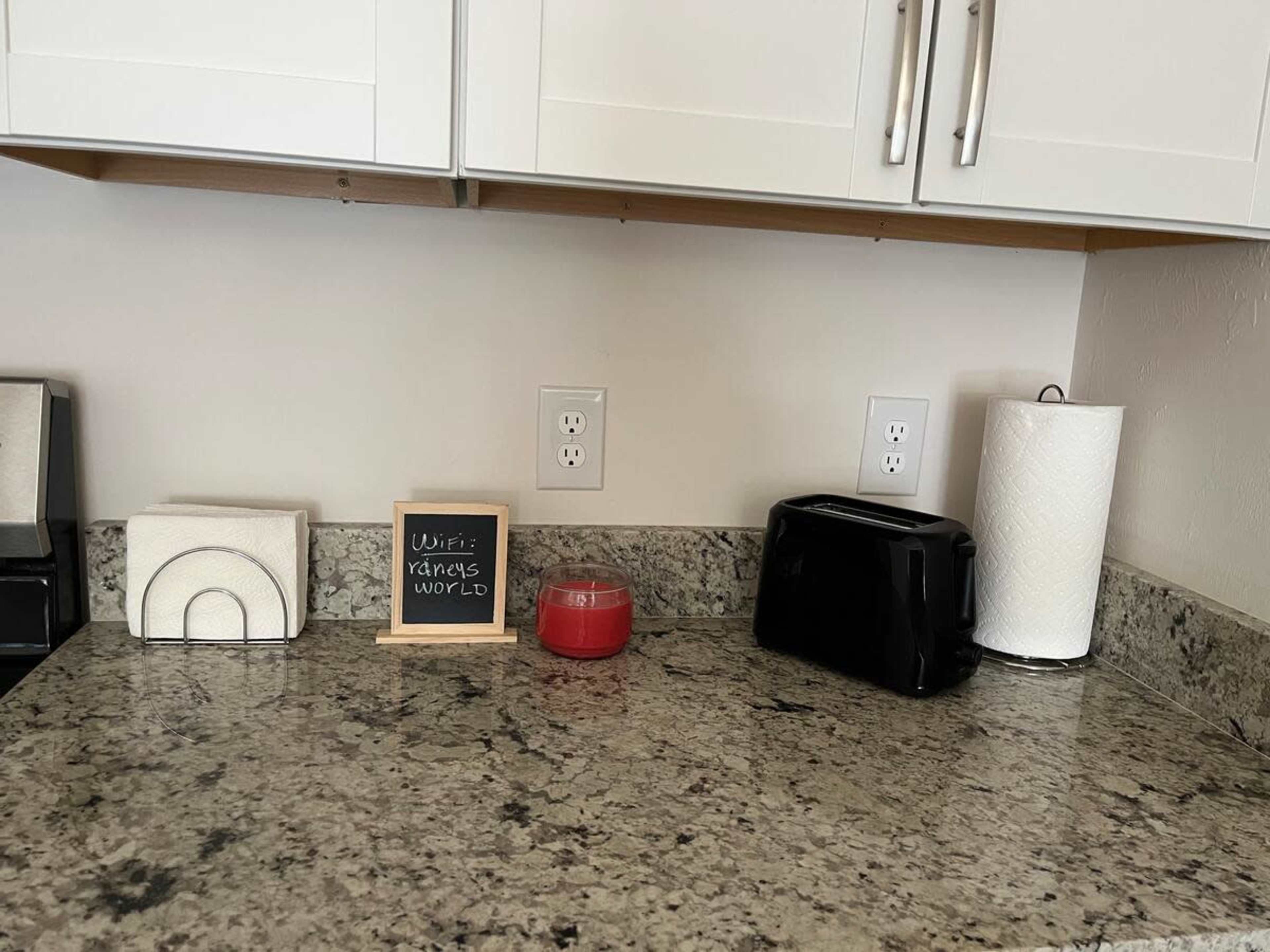 A kitchen countertop with a chalkboard, a red candle, a black toaster, and a roll of paper towels.
