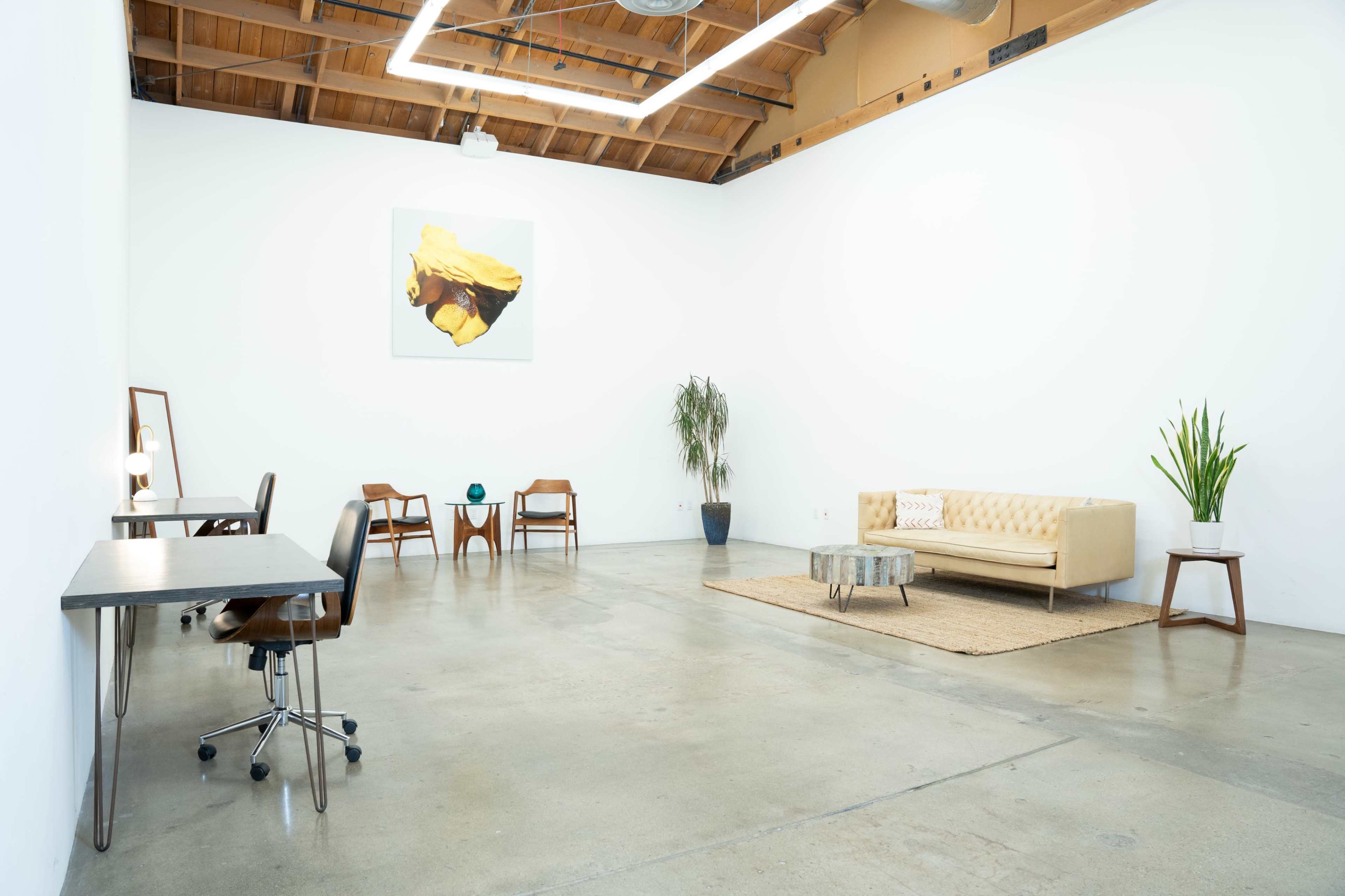 A spacious, minimalist indoor area with a sofa, a coffee table, two desk setups, and a potted plant, all arranged on a concrete floor.