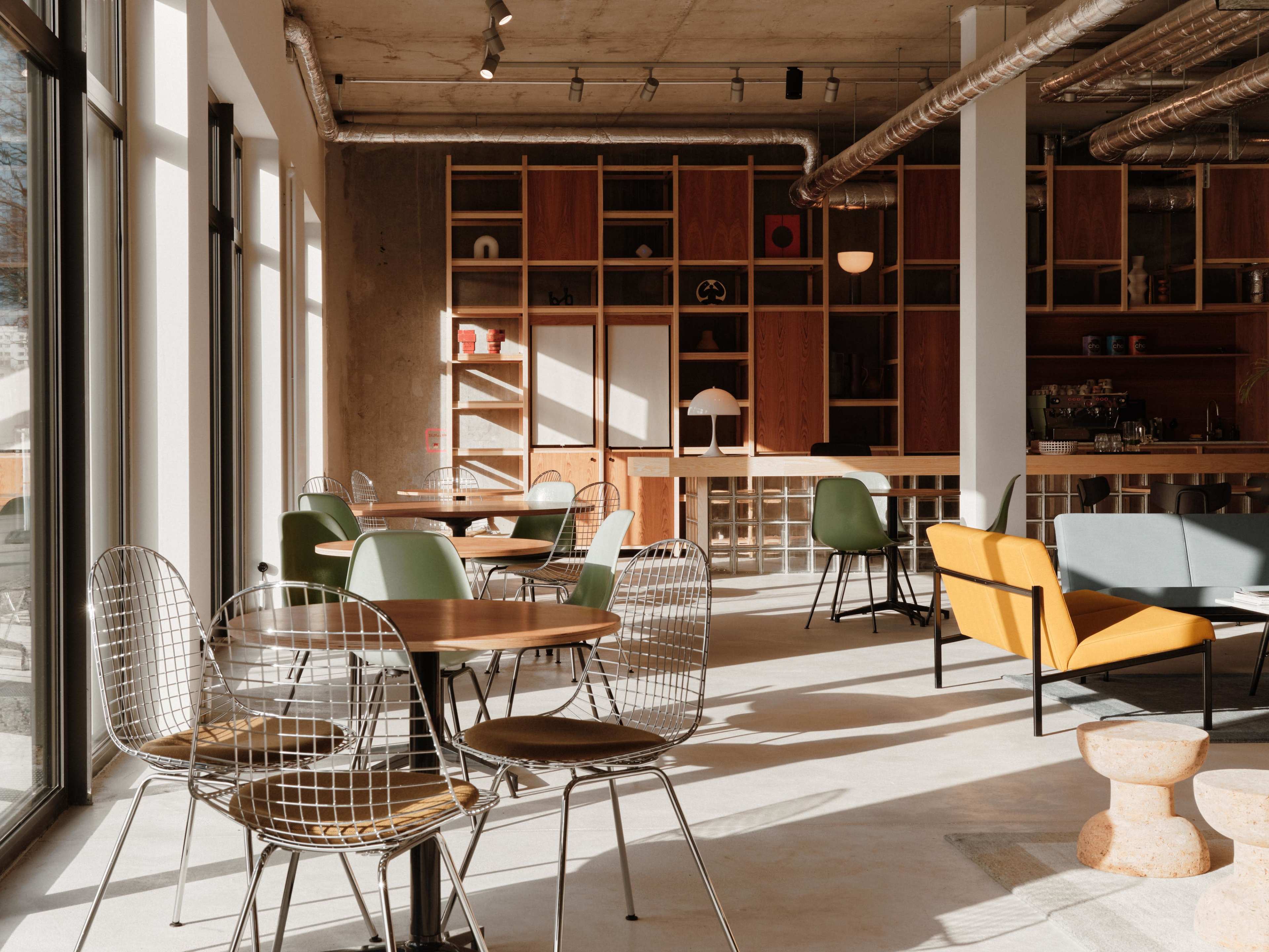 The image shows a modern café interior with various seating arrangements, large windows allowing natural light, and a wooden bar area in the background.