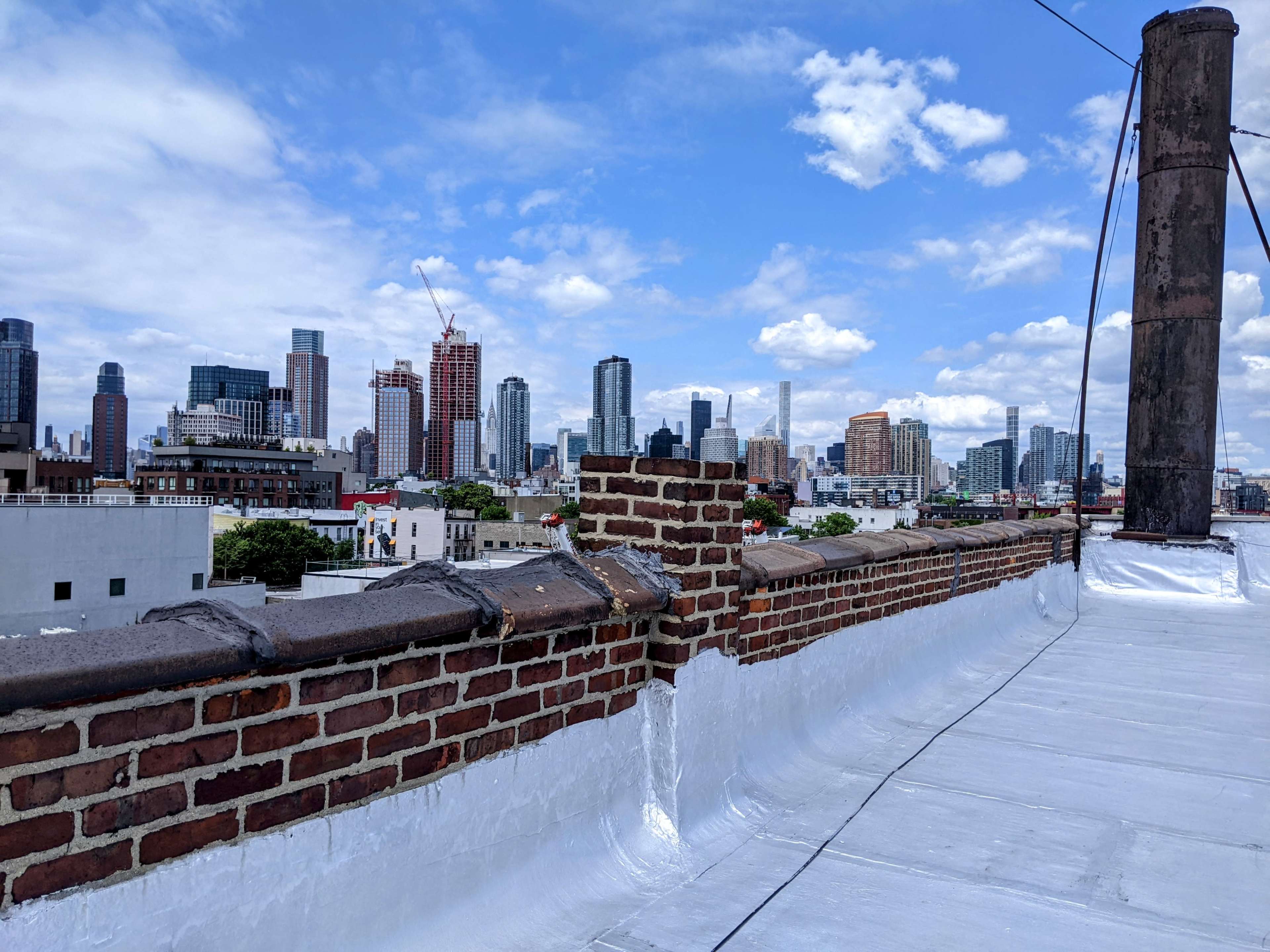 A view from a roof featuring a brick parapet and a skyline of tall buildings under a partly cloudy sky.