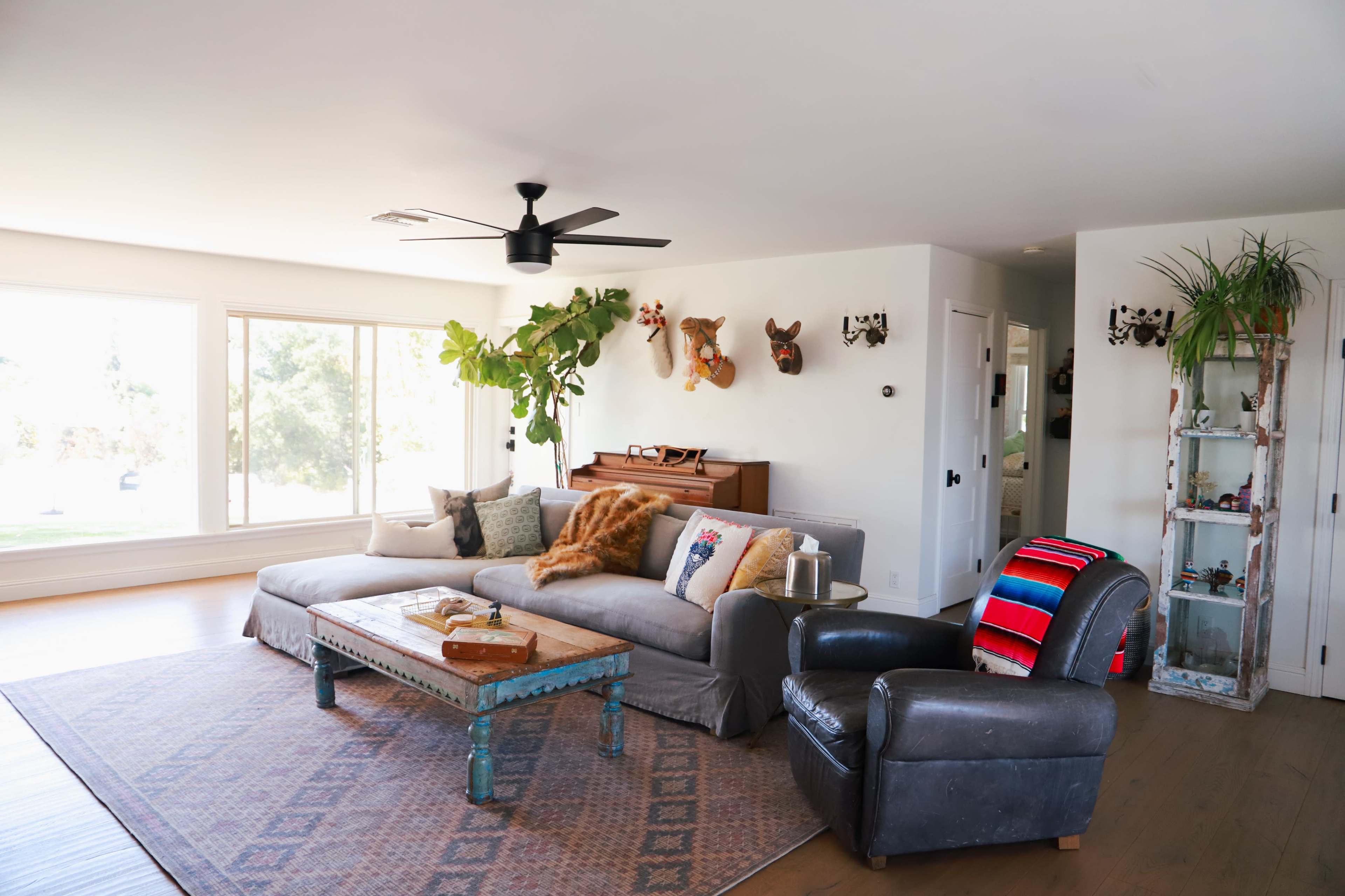 The image shows a modern living room featuring a large sectional sofa, a wooden coffee table, and a plush armchair, with decorative plants and artwork on the walls.