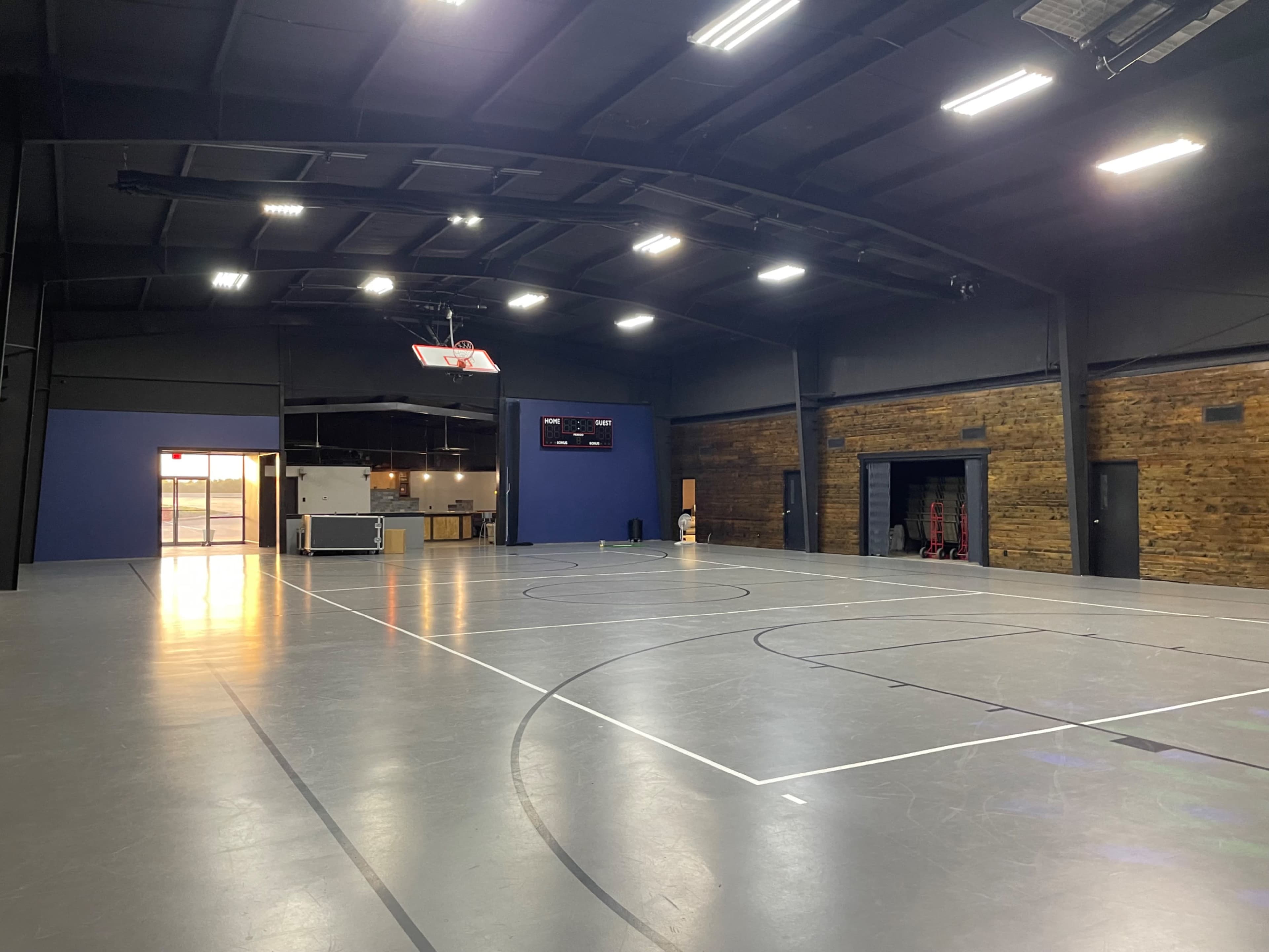 The image shows an empty indoor gymnasium with polished gray floors, basketball hoops, and a wooden accent wall.