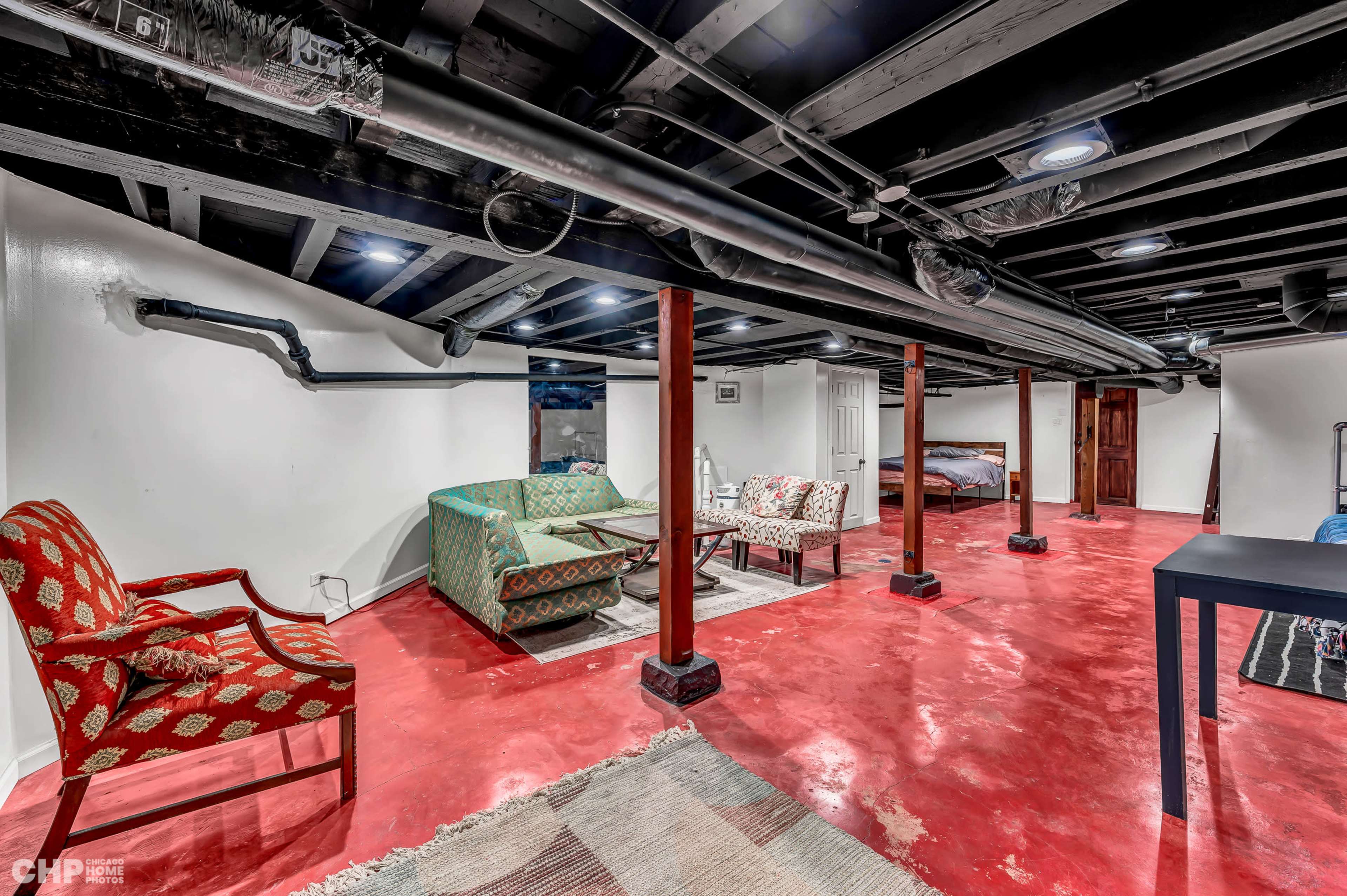 A finished basement with a red concrete floor, two sofas, and several furnishings arranged beneath black beams and pipes.