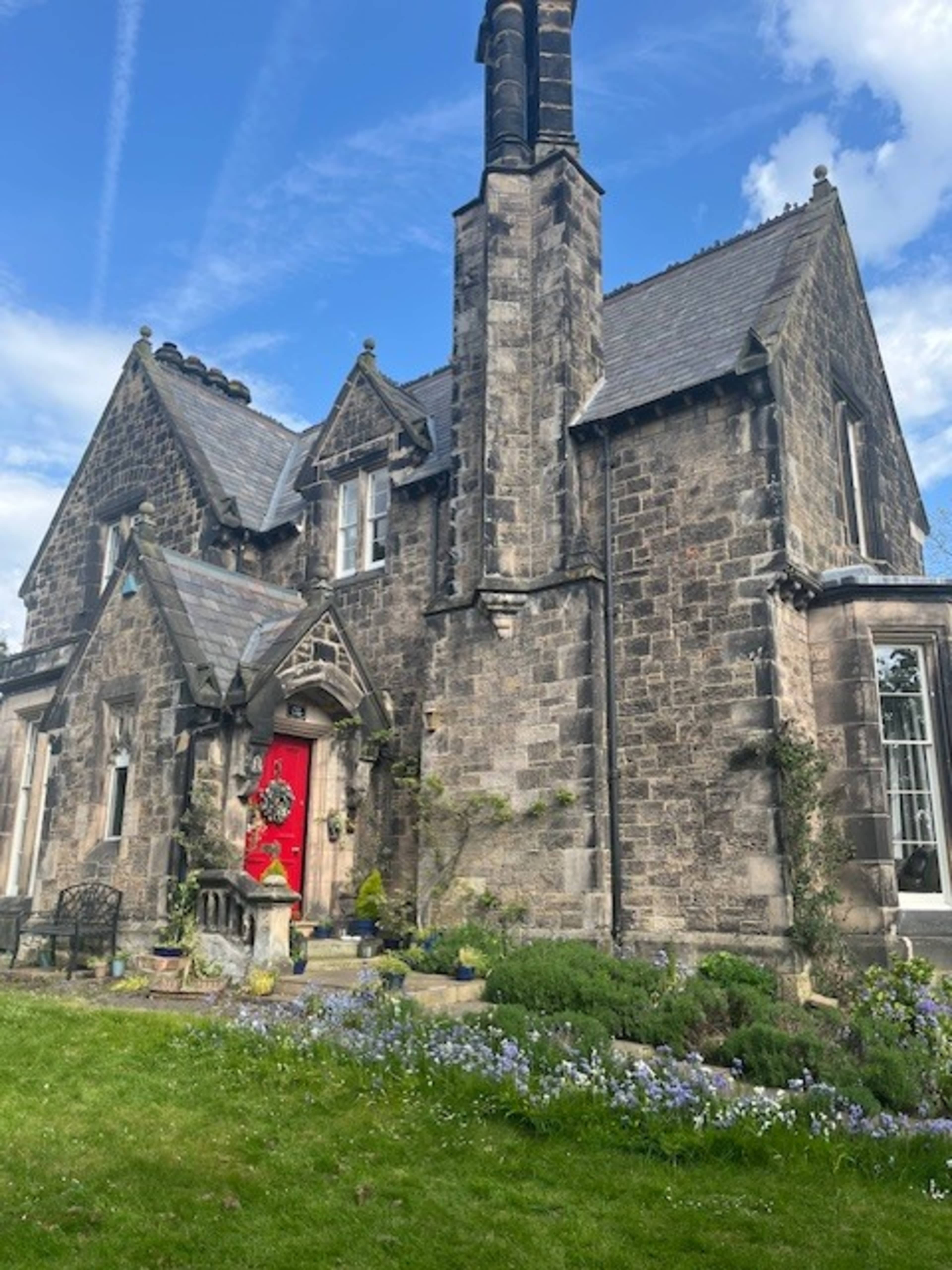 A stone house featuring a prominent red door and surrounded by a garden with blue flowers.