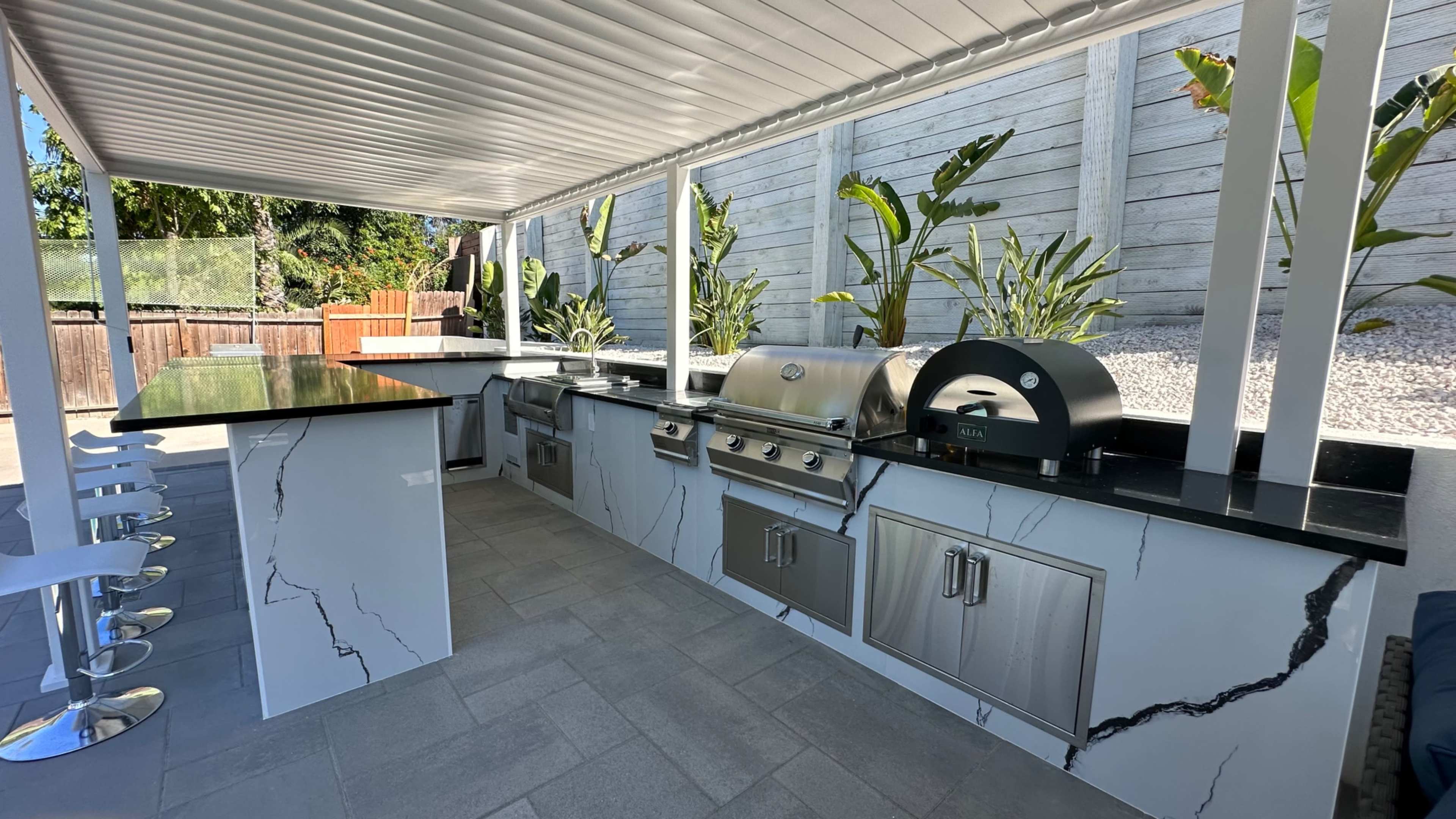 An outdoor kitchen area featuring stainless steel appliances, a black and white marble countertop, and a pizza oven, all shaded by a roof with vertical supports.