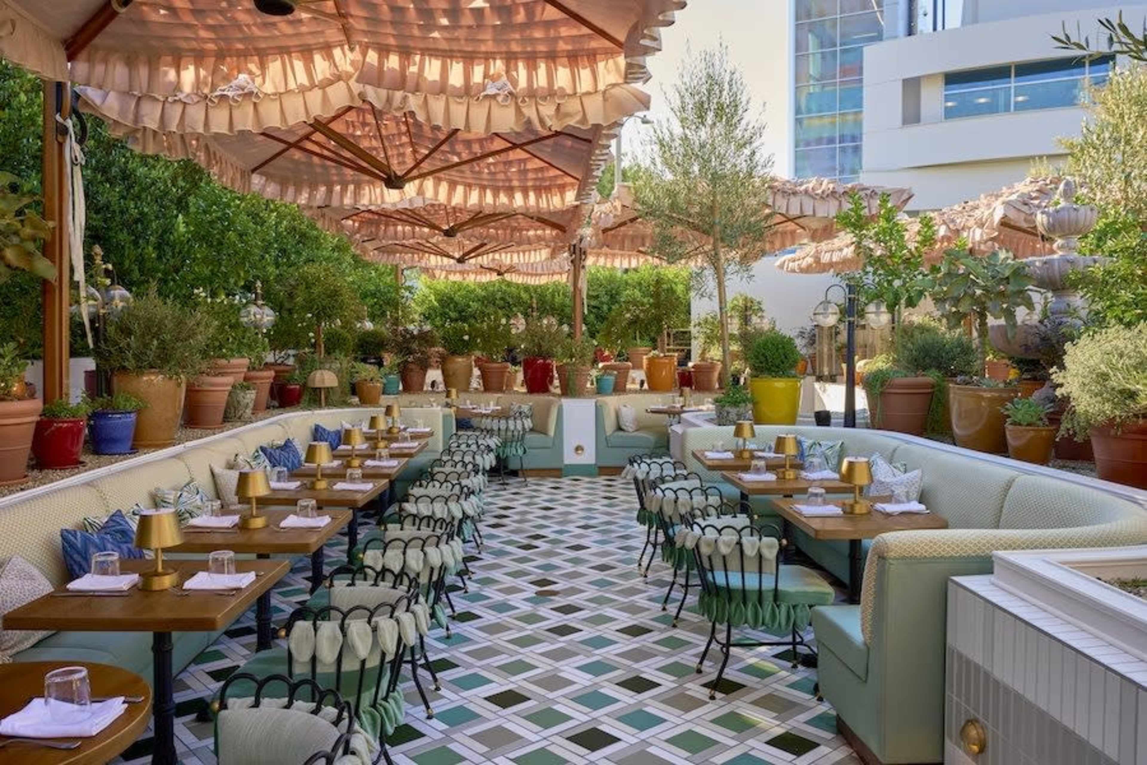 The image shows an outdoor restaurant patio decorated with potted plants, tables set with tableware, and large umbrellas for shade.