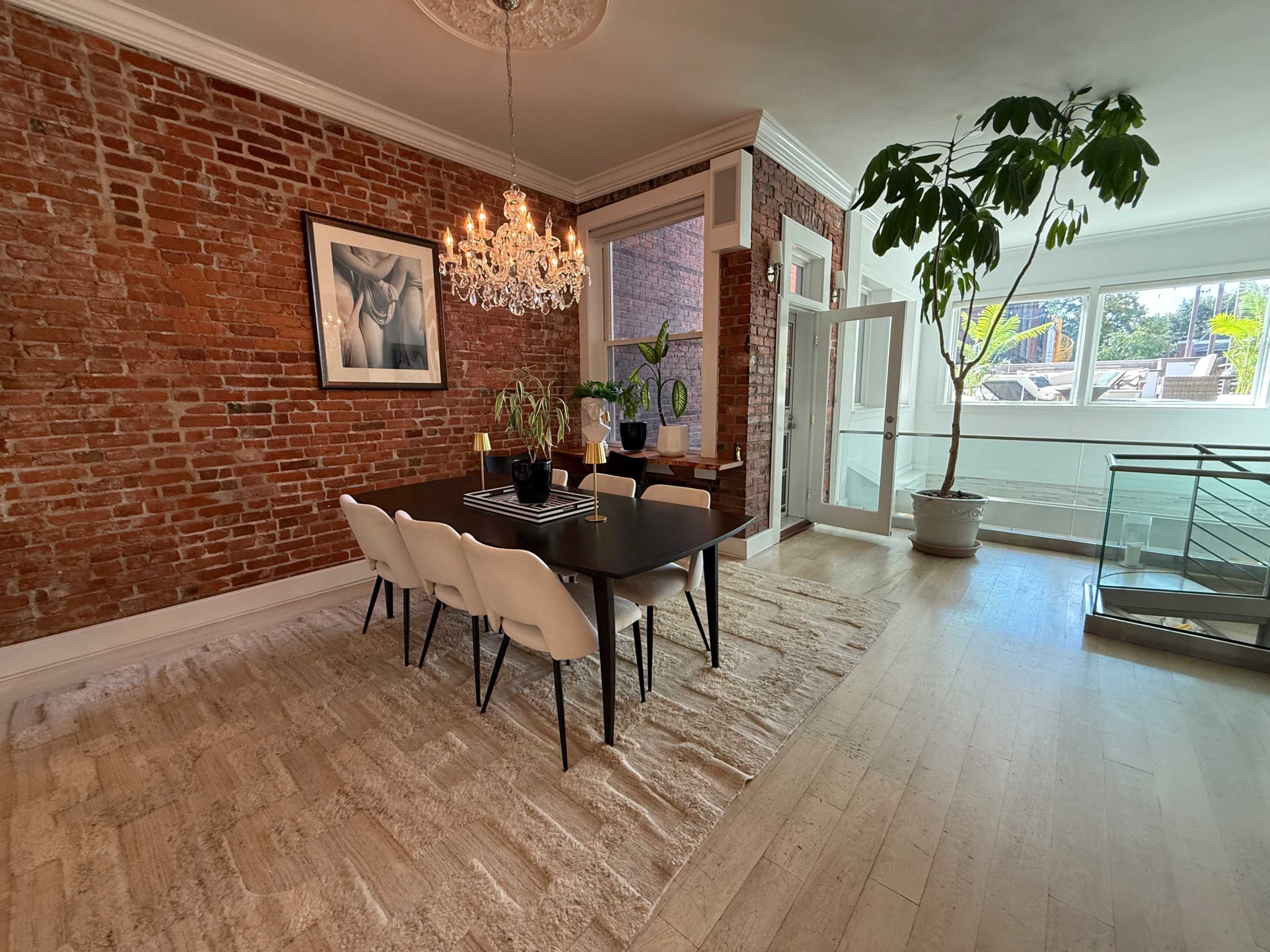 The image shows a dining area with a black table surrounded by white chairs, set against a backdrop of exposed brick walls and illuminated by a chandelier.