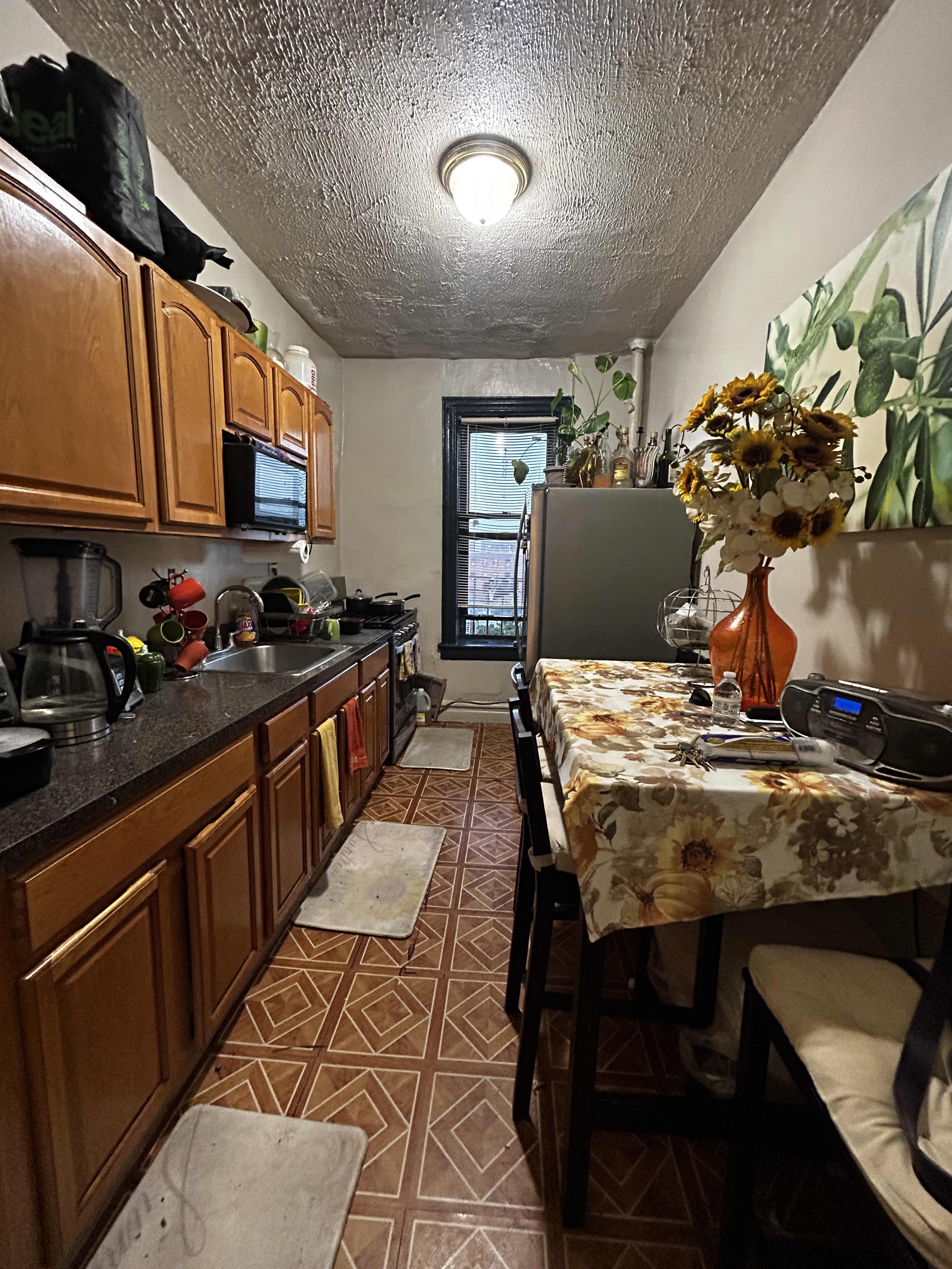 A narrow kitchen features wooden cabinets, a table set with a floral tablecloth, and appliances along the walls.