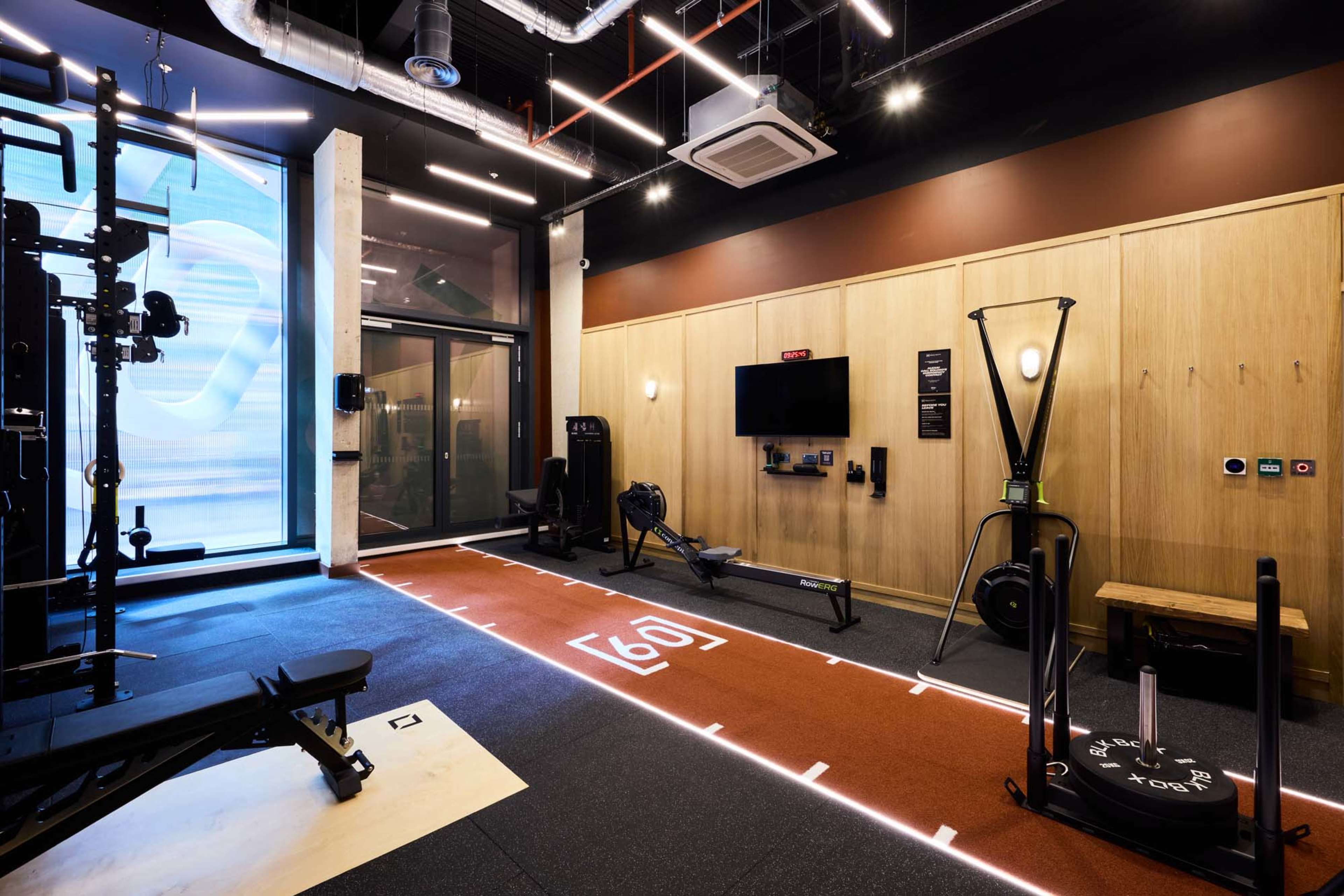 The image shows a modern gym interior equipped with a variety of fitness equipment, including a weight bench, rowing machine, and resistance machines, alongside a large window allowing natural light to enter.