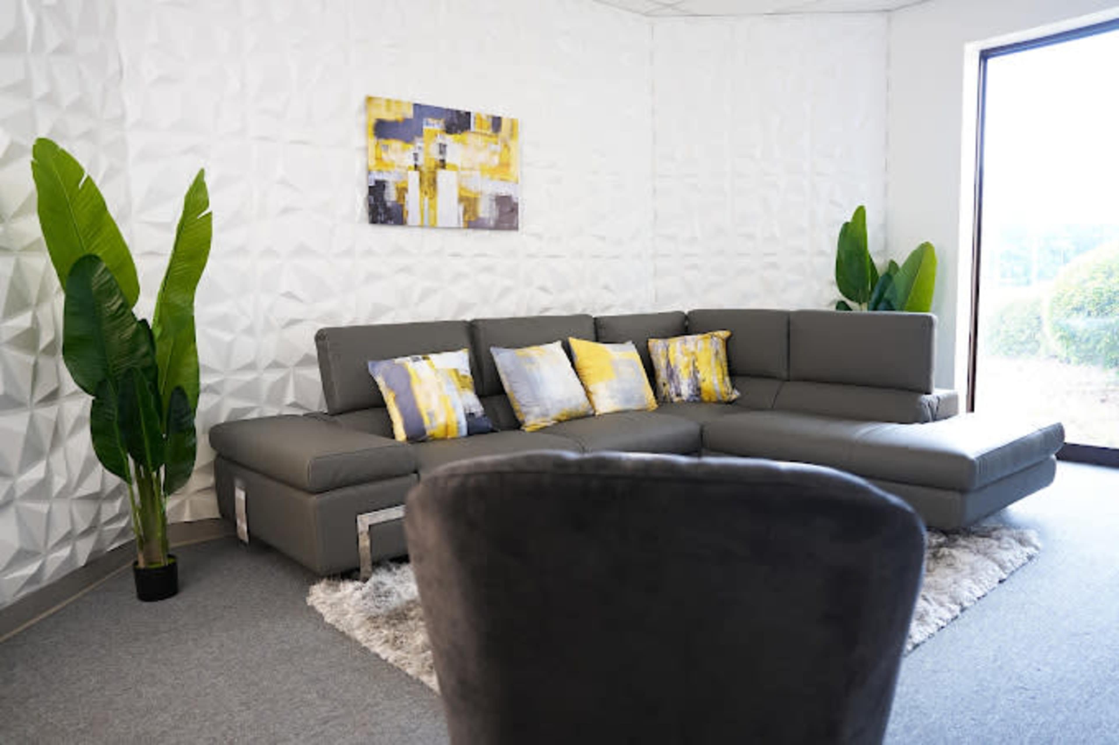 The image shows a modern gray sectional sofa with decorative pillows, positioned in a well-lit room featuring textured white walls and potted plants.