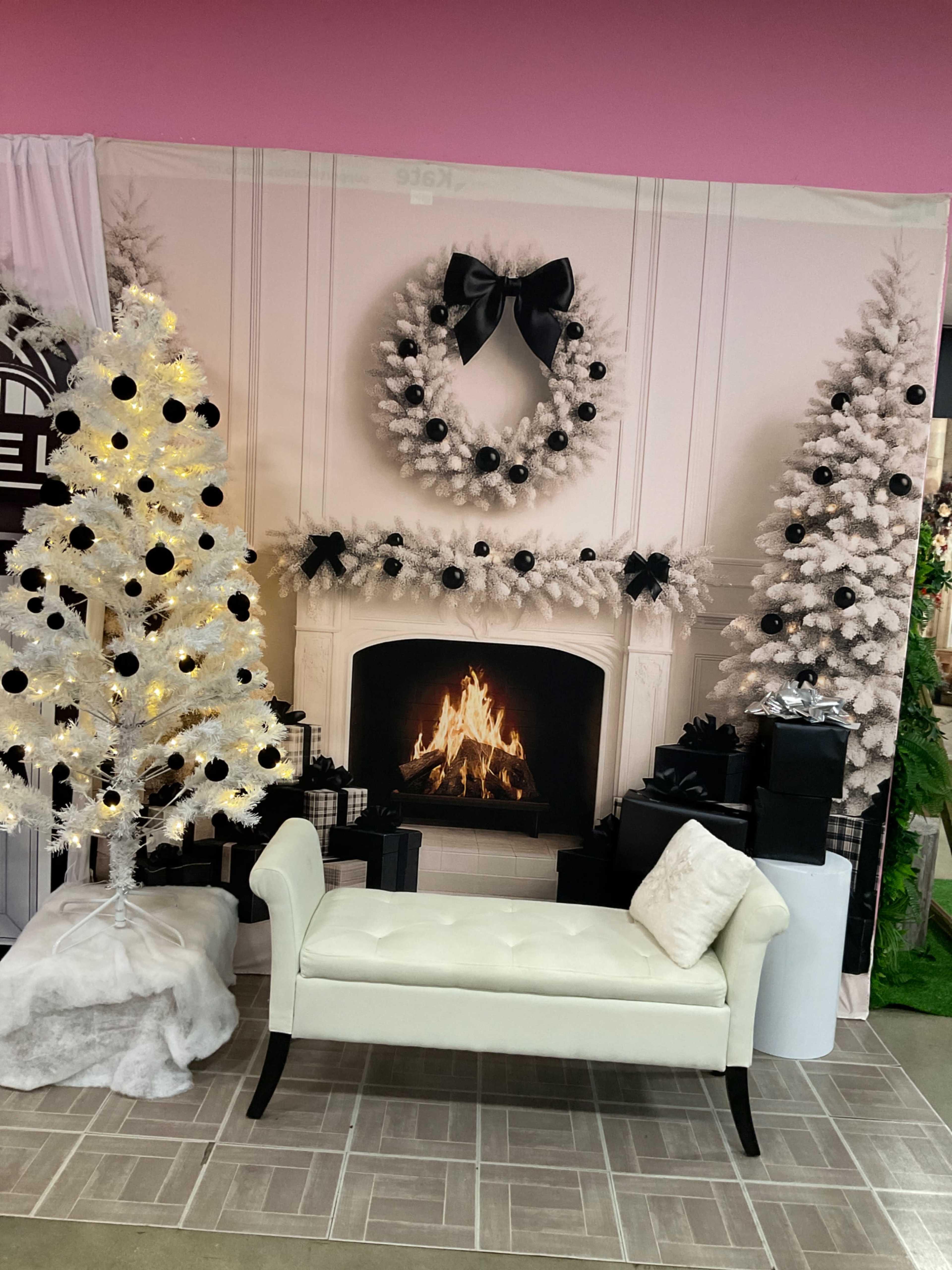 The image shows a festive display with a fireplace flanked by two decorated Christmas trees, a white bench in front, and several wrapped presents nearby.
