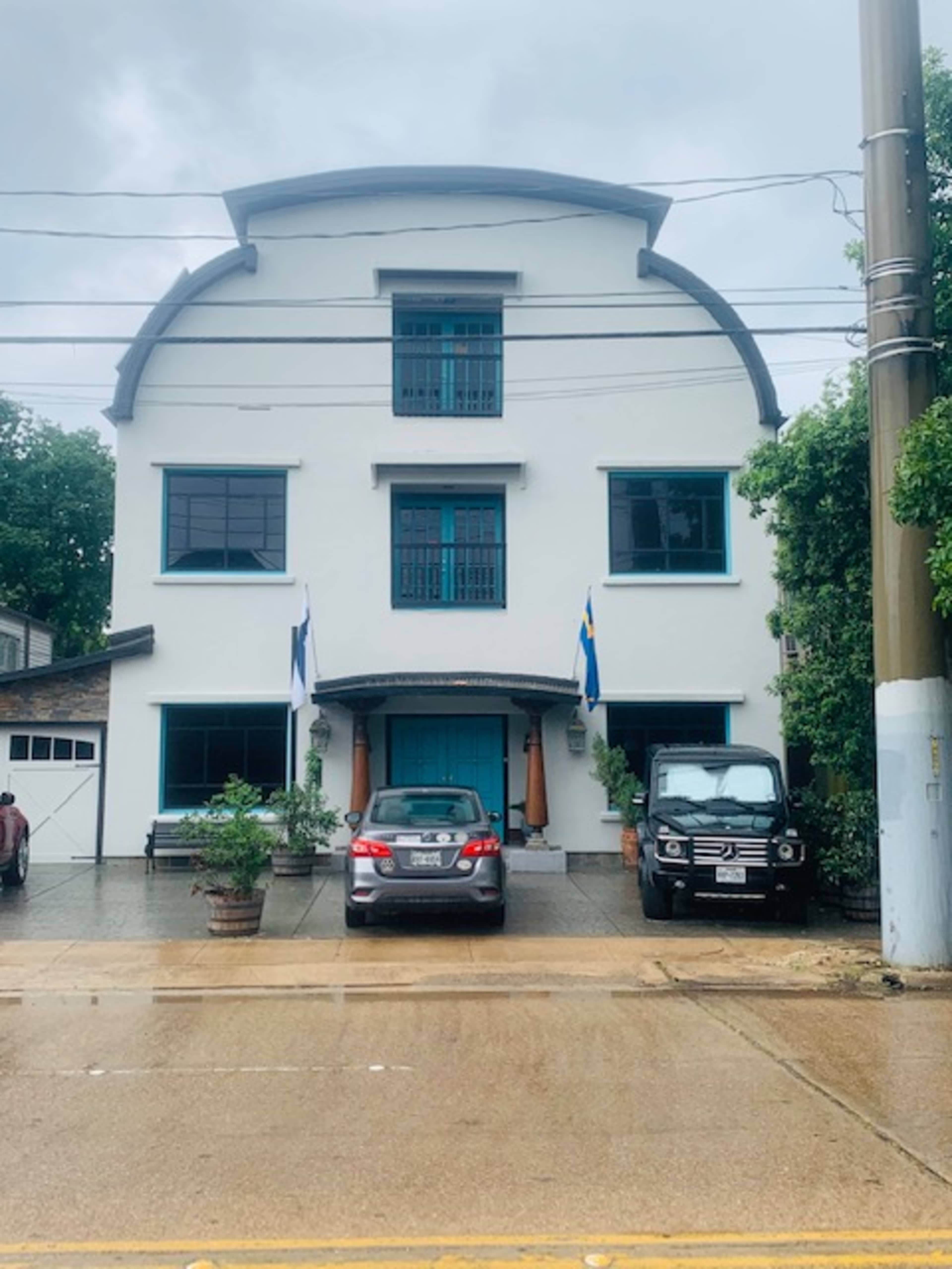 The image shows a white two-story building with a curved roof, featuring blue windows and parked cars in front.