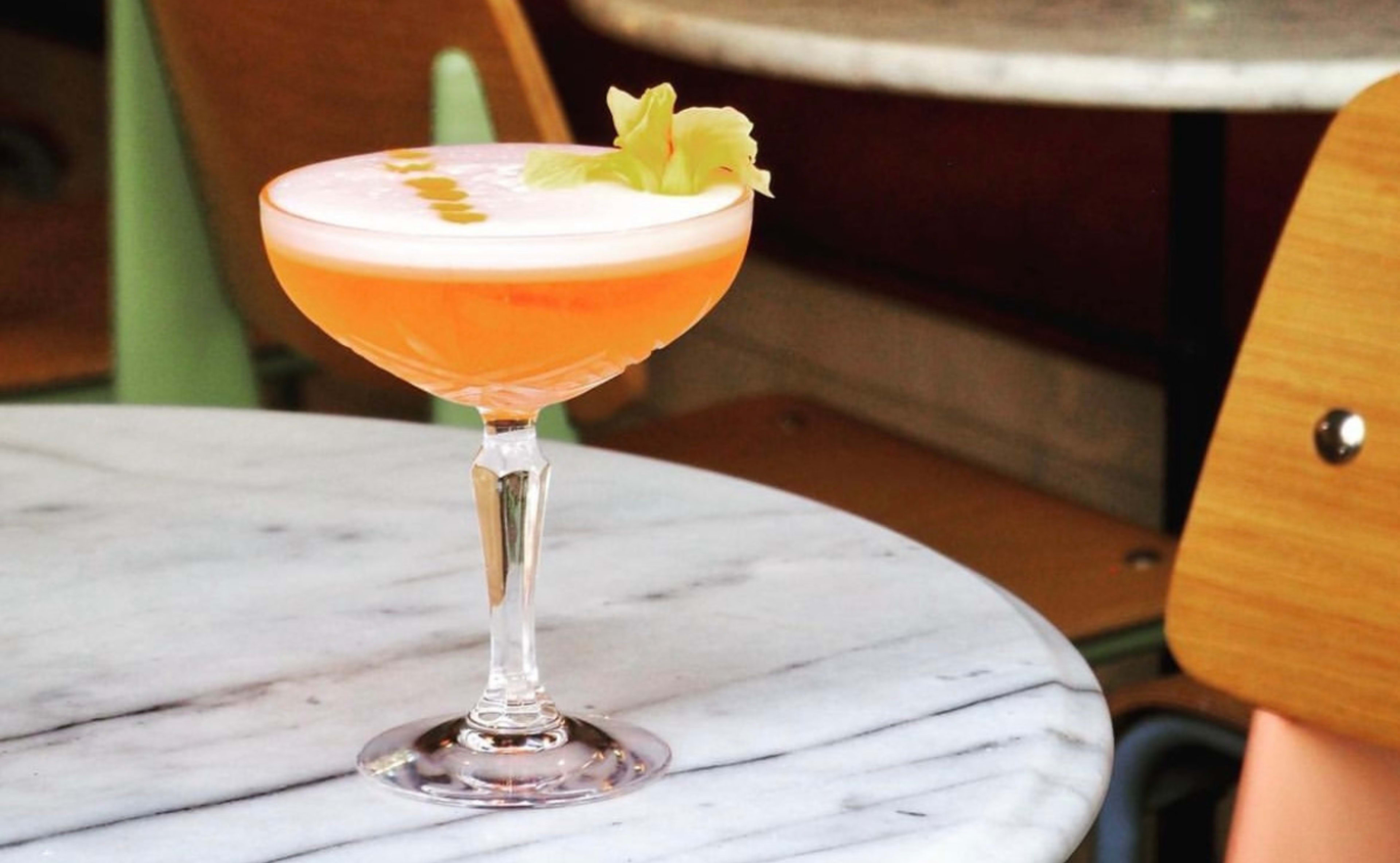 A cocktail in a glass garnished with a flower sits on a marble table next to wooden chairs.